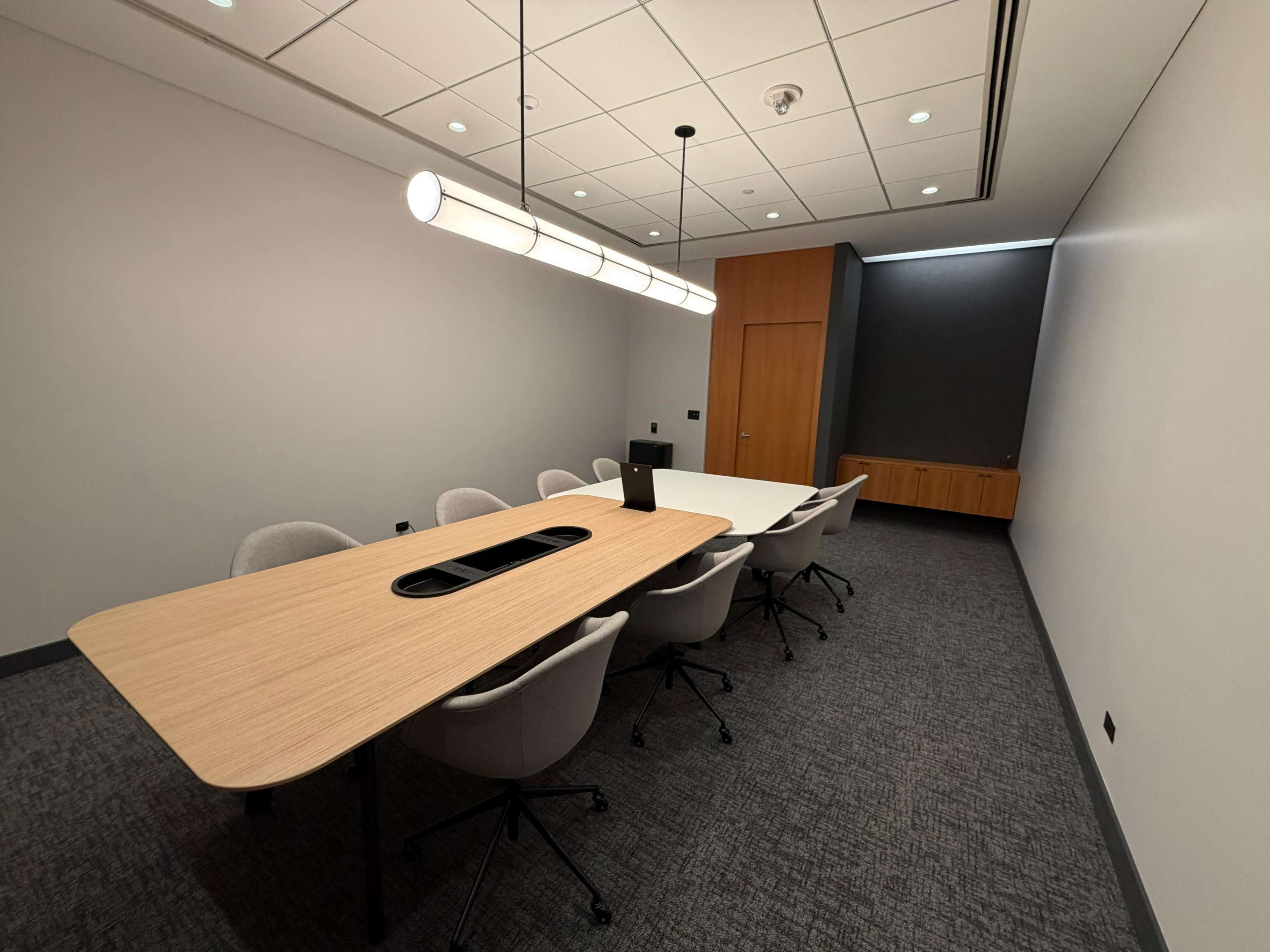 The image shows a modern conference room with a long wooden table surrounded by ergonomic chairs and a wall-mounted display.