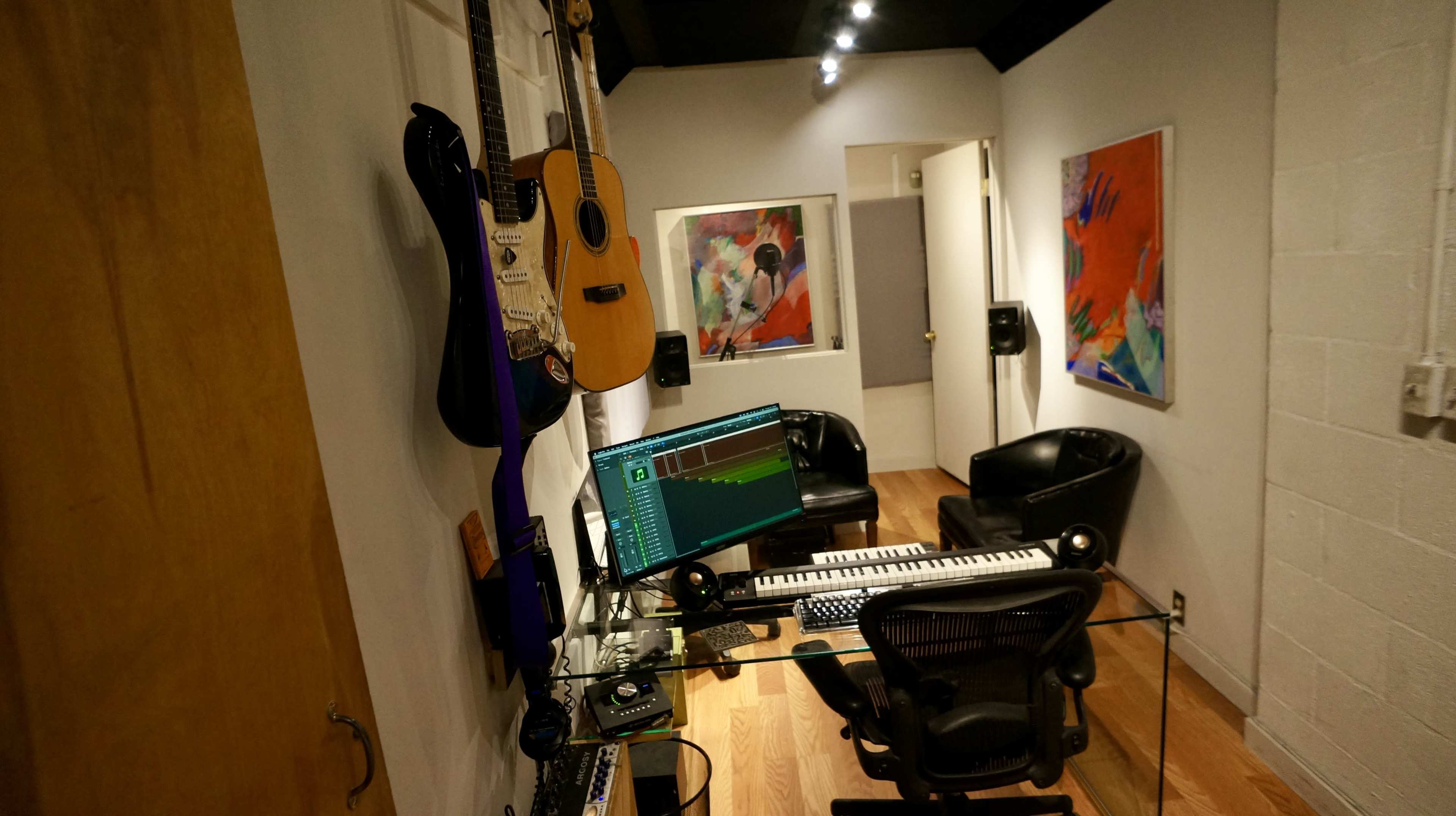 The image shows a small music studio featuring a keyboard, computer, and guitars on the wall, with two black chairs and colorful artwork on display.