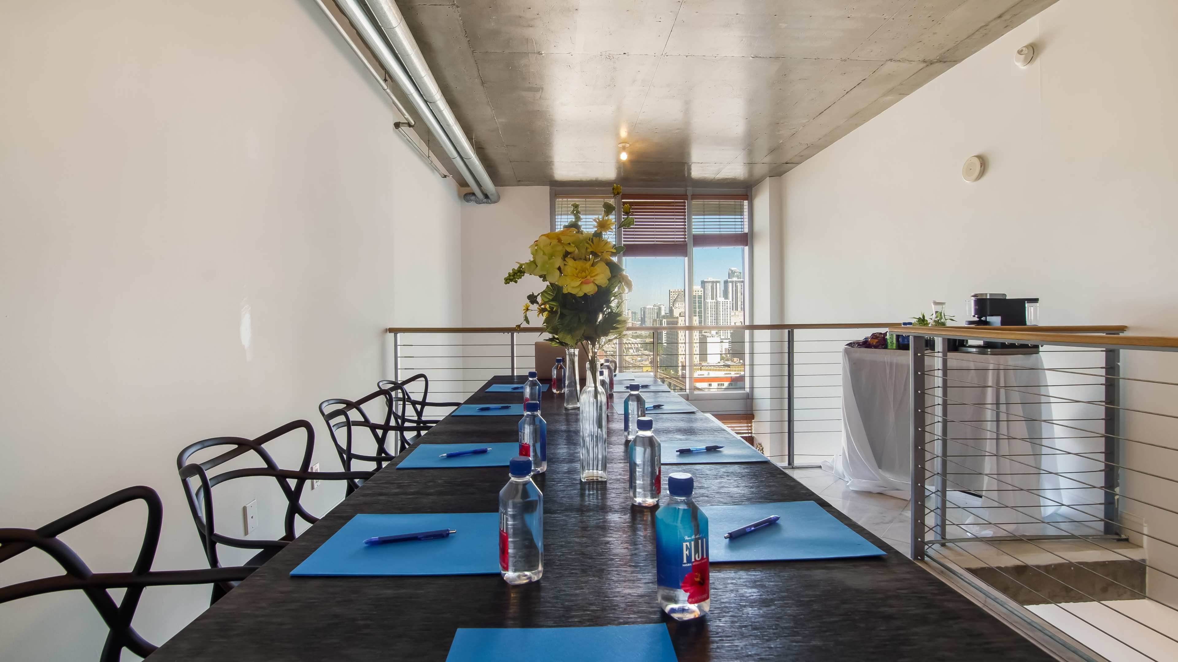 A long conference table is set with blue notepads and bottles of water, featuring a floral centerpiece, against a backdrop of city skyline views from a modern office space.