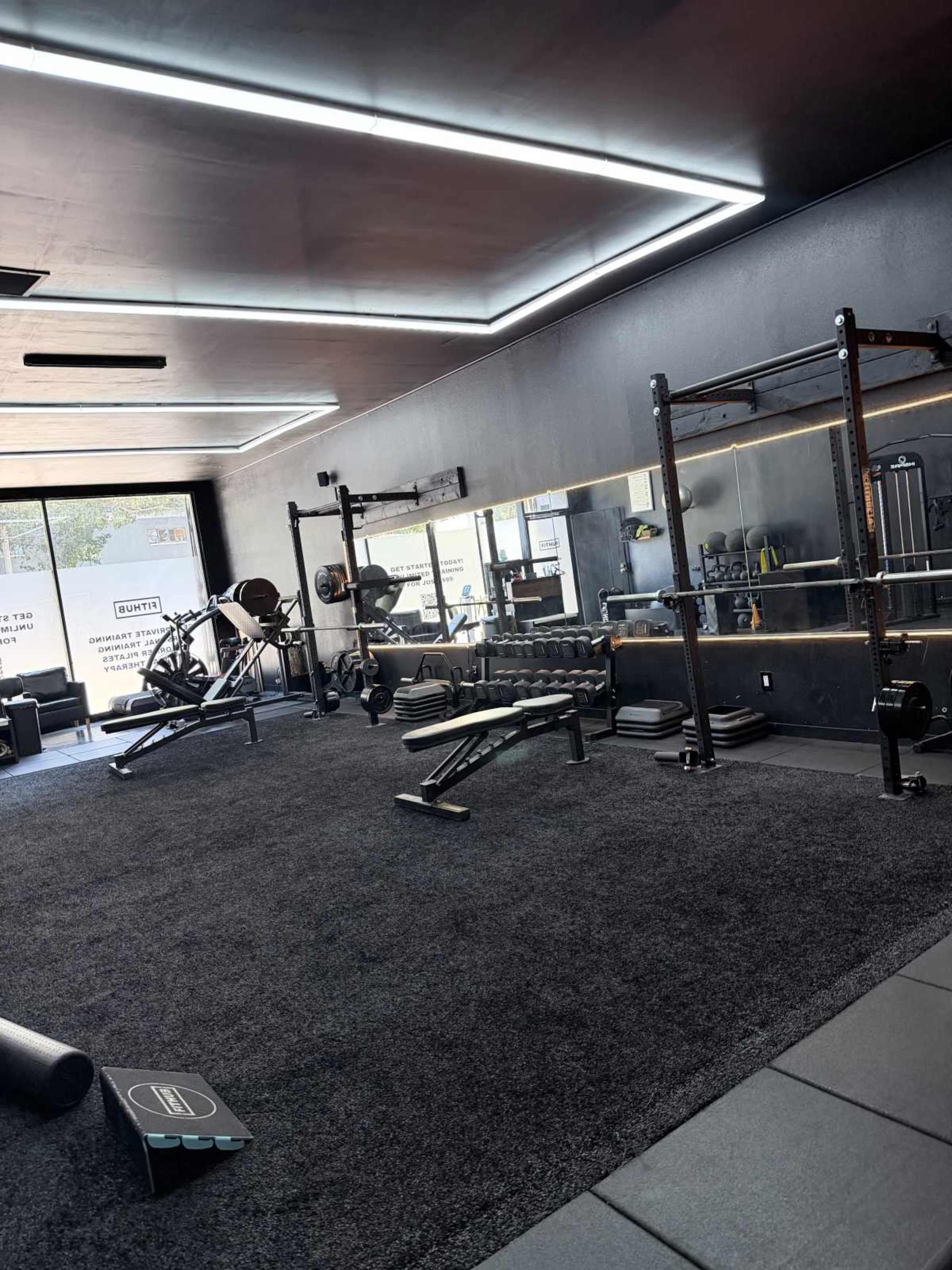 The image shows a modern gym interior featuring various exercise equipment, including weights, benches, and a mirror, all set on a carpeted floor.