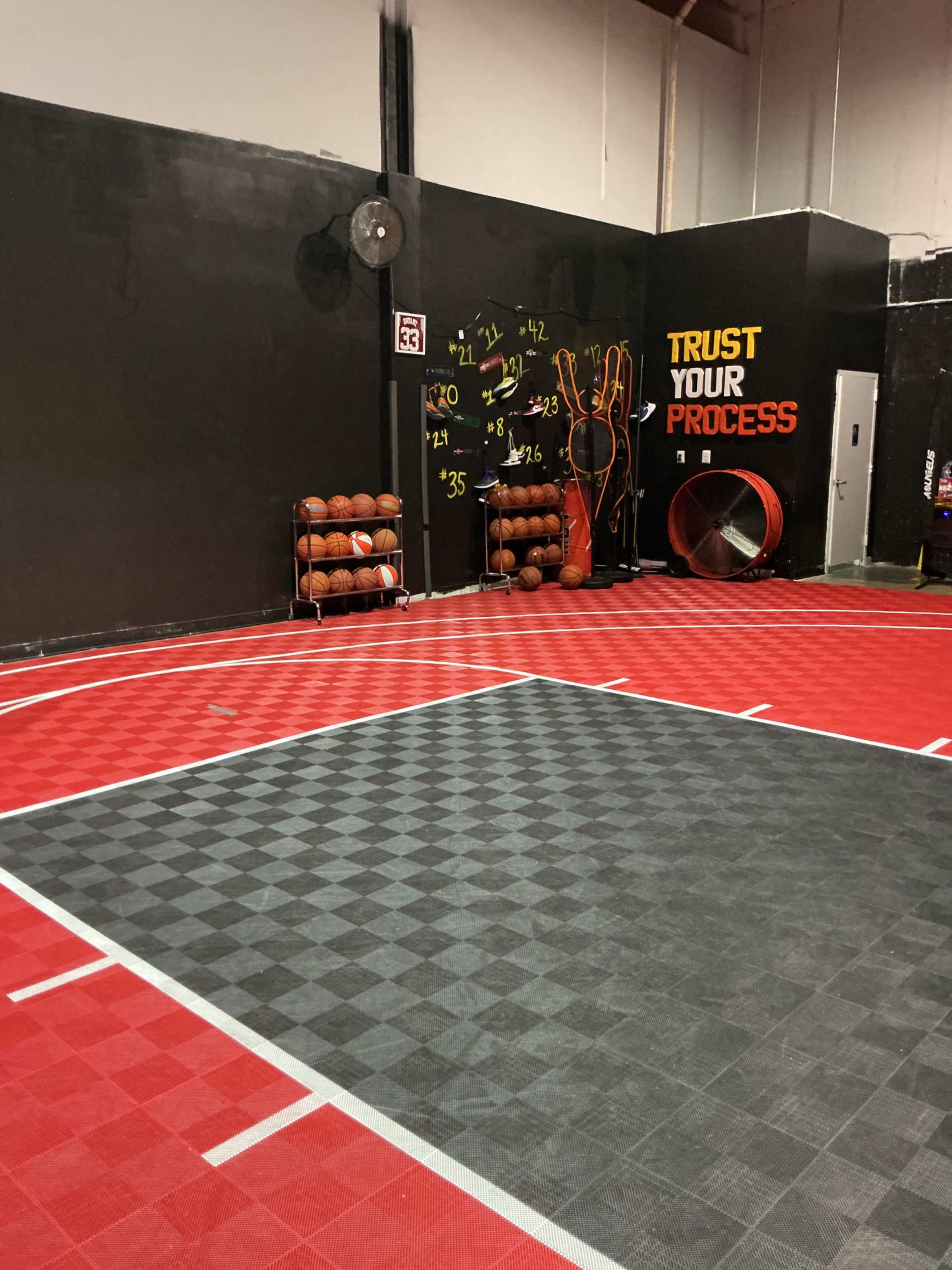 The image shows a basketball training facility with a red and black checkered court, a wall displaying the words "TRUST YOUR PROCESS," and several basketballs stacked on a rack.