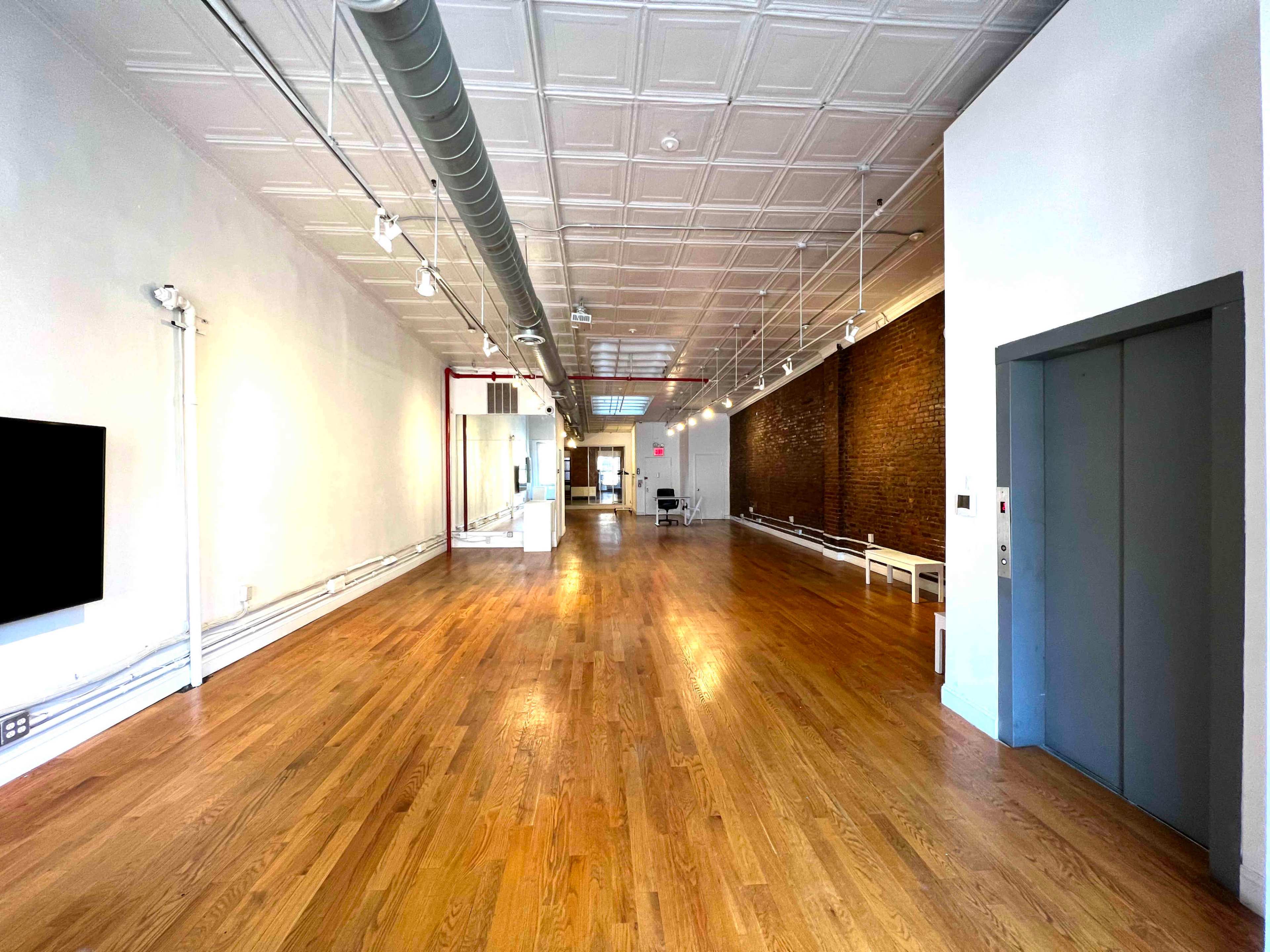 The image shows a spacious, well-lit interior with hardwood flooring, exposed brick walls, and an elevator on one side.