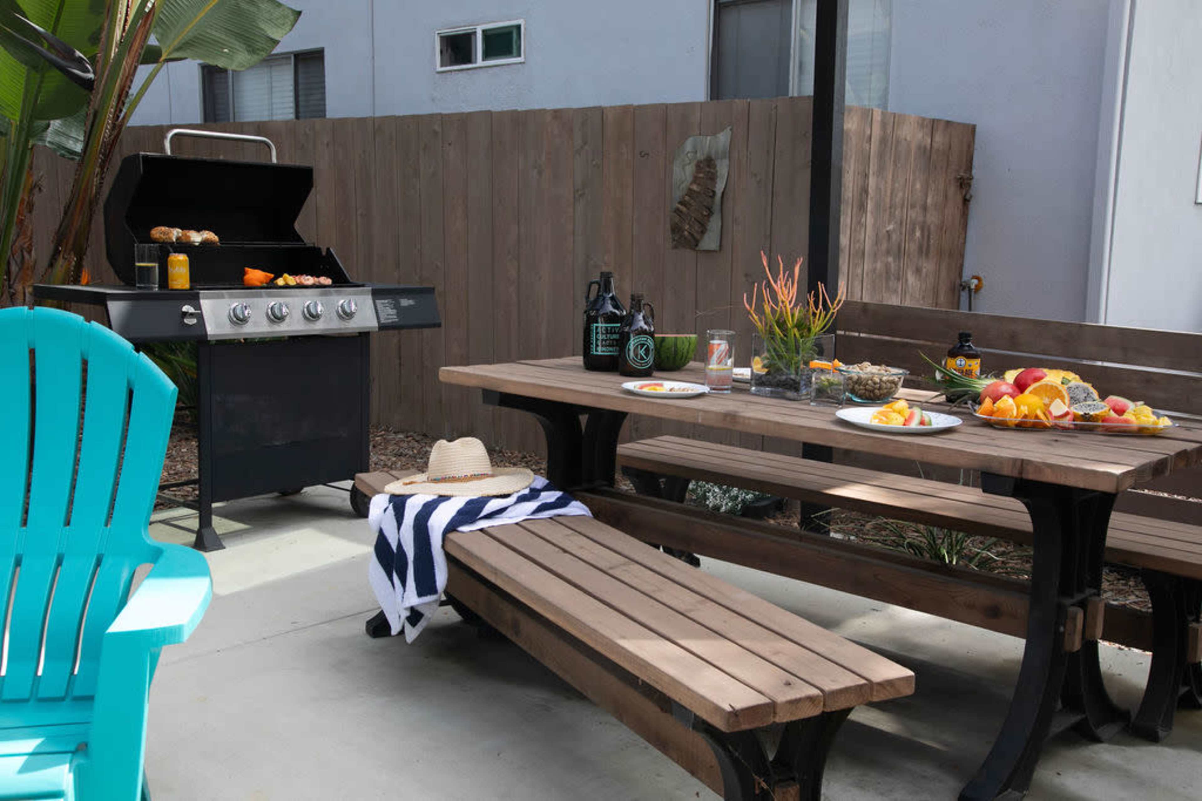 The scene features a backyard patio with a barbecue grill, a wooden dining table set with food, and an Adirondack chair nearby.