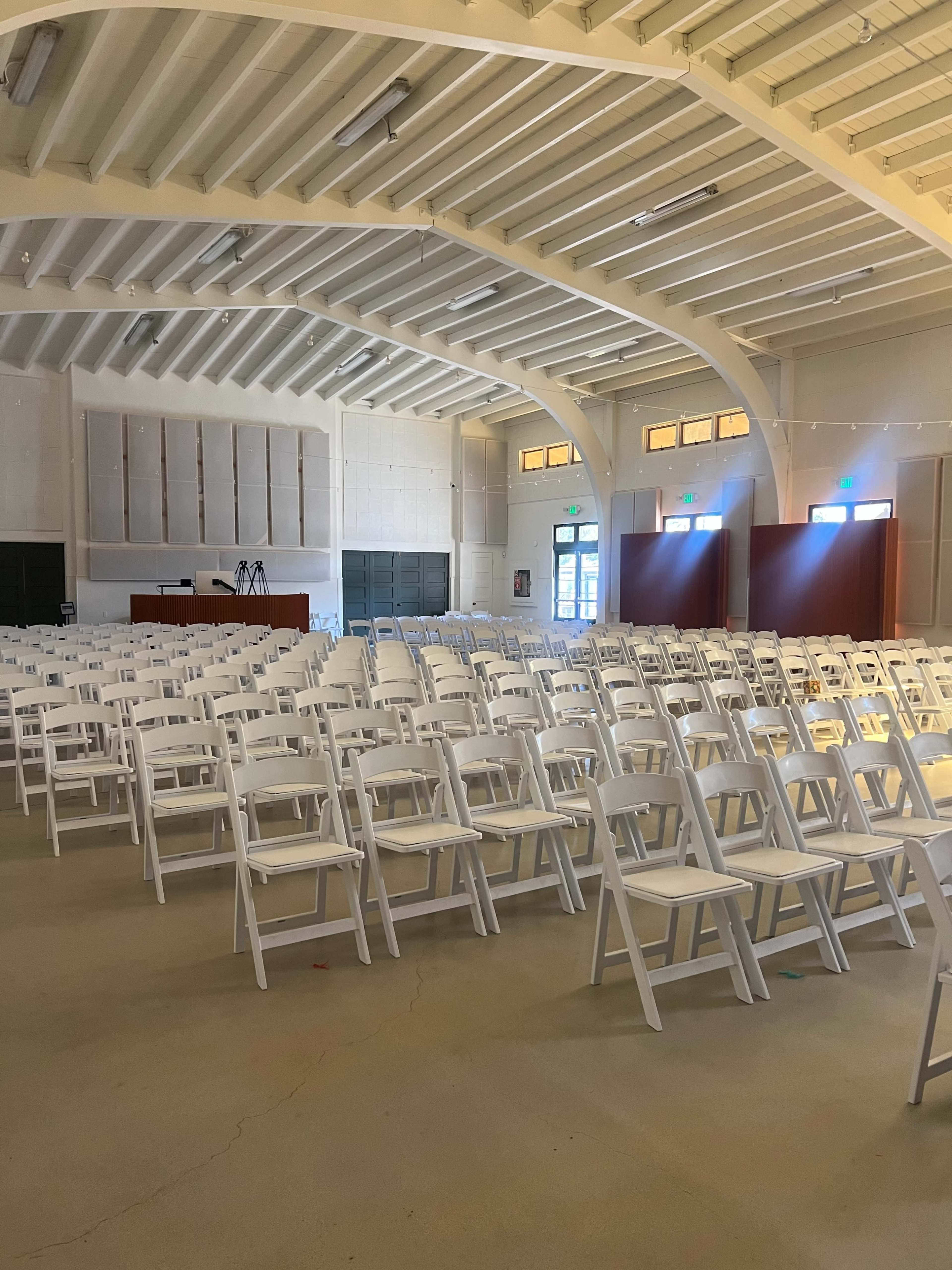 The image shows a large indoor space with rows of white folding chairs set up facing a stage area at the front.