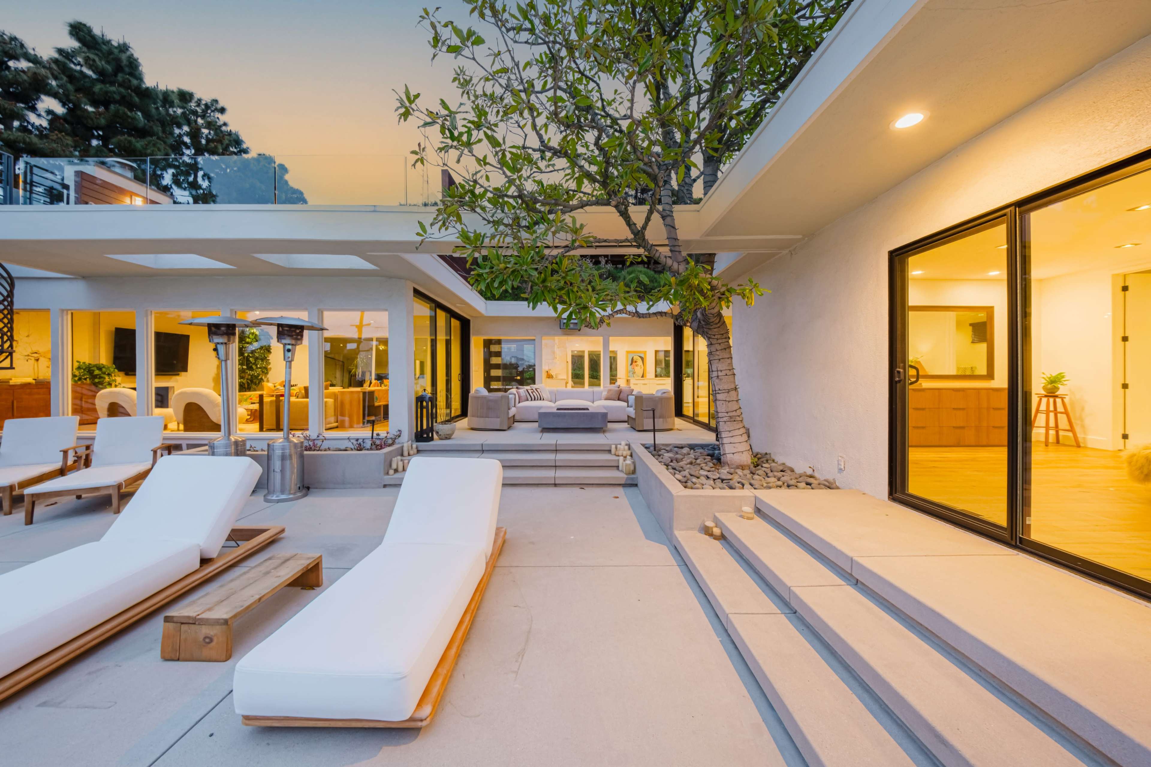 A modern outdoor patio features lounge chairs, a table, and large sliding glass doors leading into an interior space with warm lighting.
