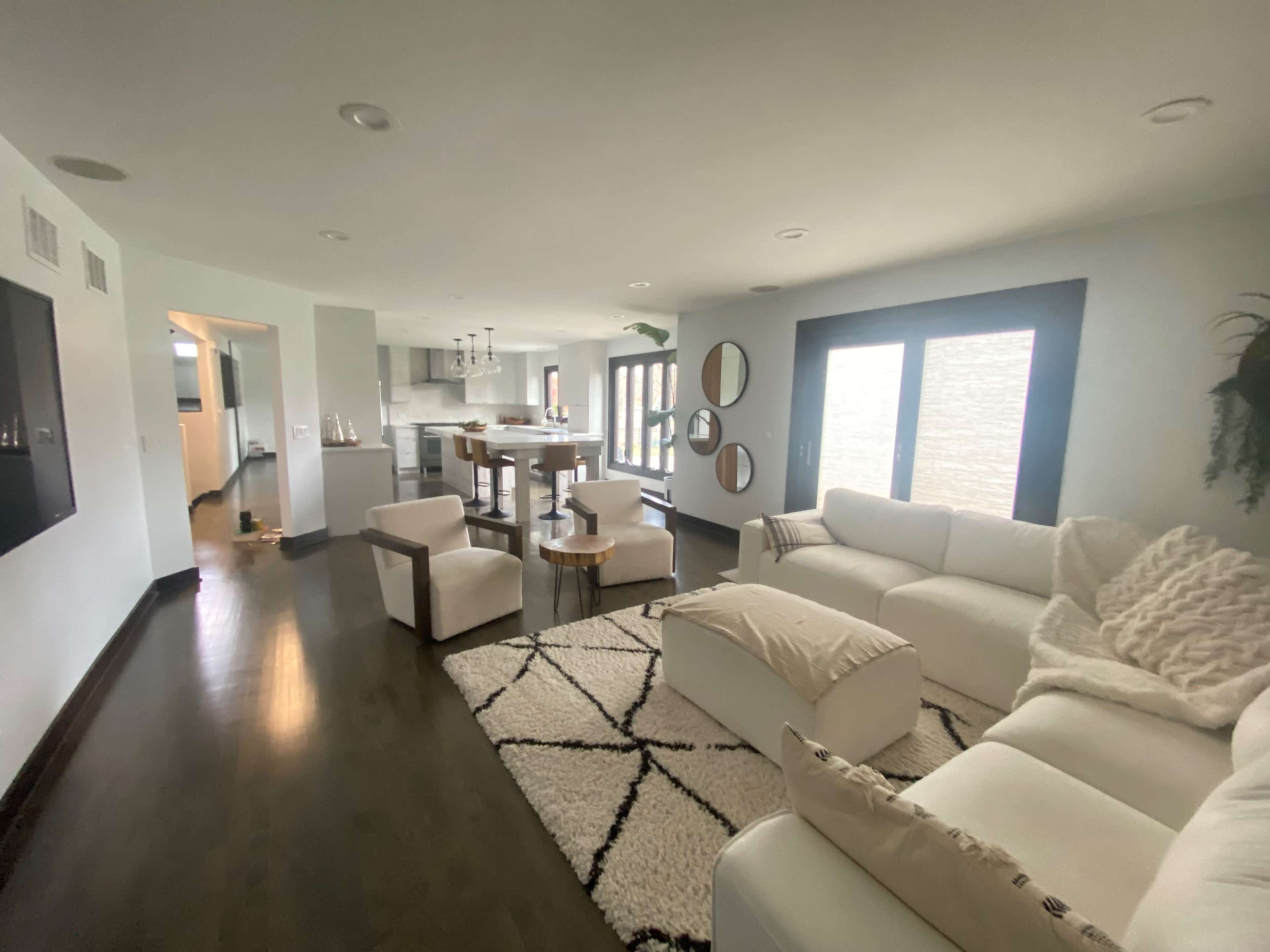 The image shows a modern, open-concept living room with a white sectional sofa, a round wooden coffee table, and large windows letting in natural light.
