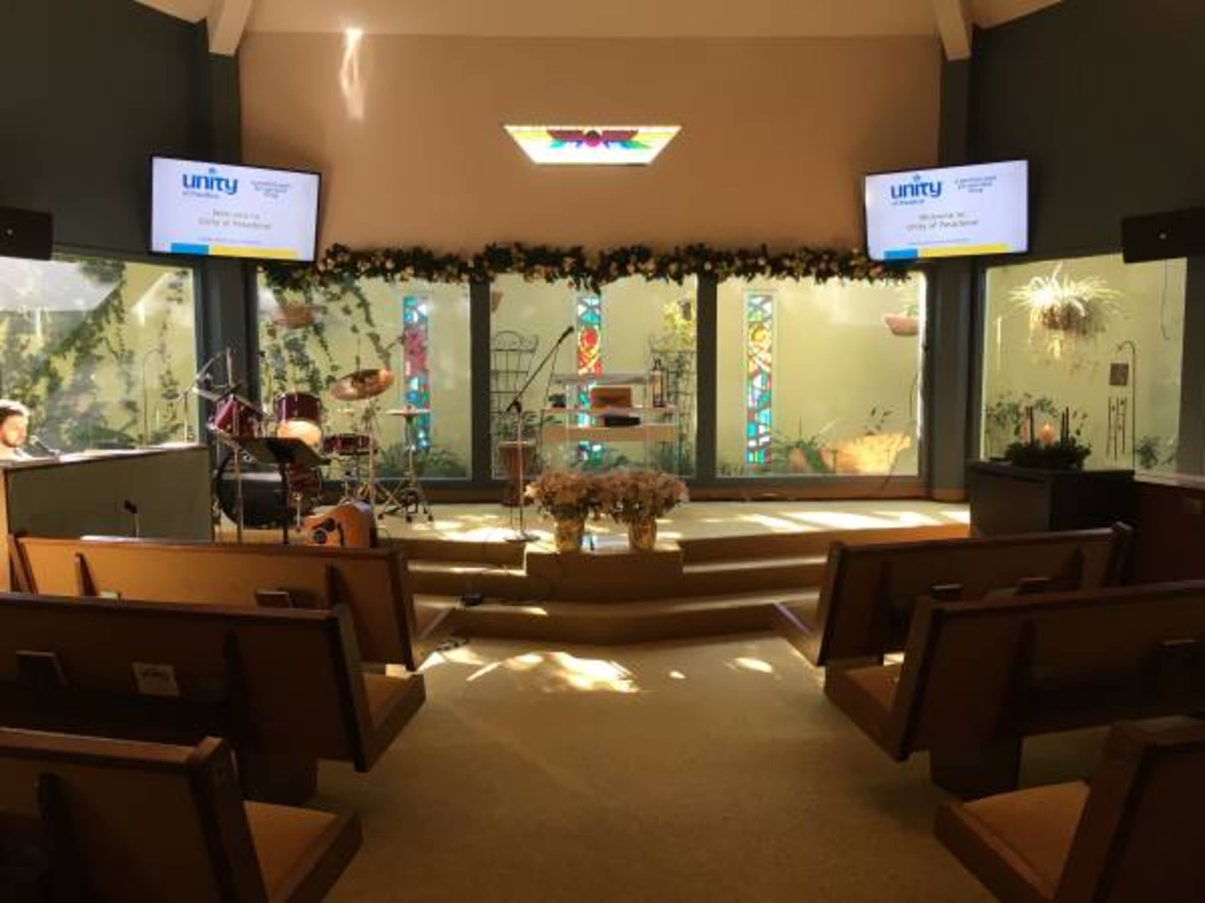 The interior of a church features a stage with musical instruments, bright stained glass windows, and seating arranged in rows.