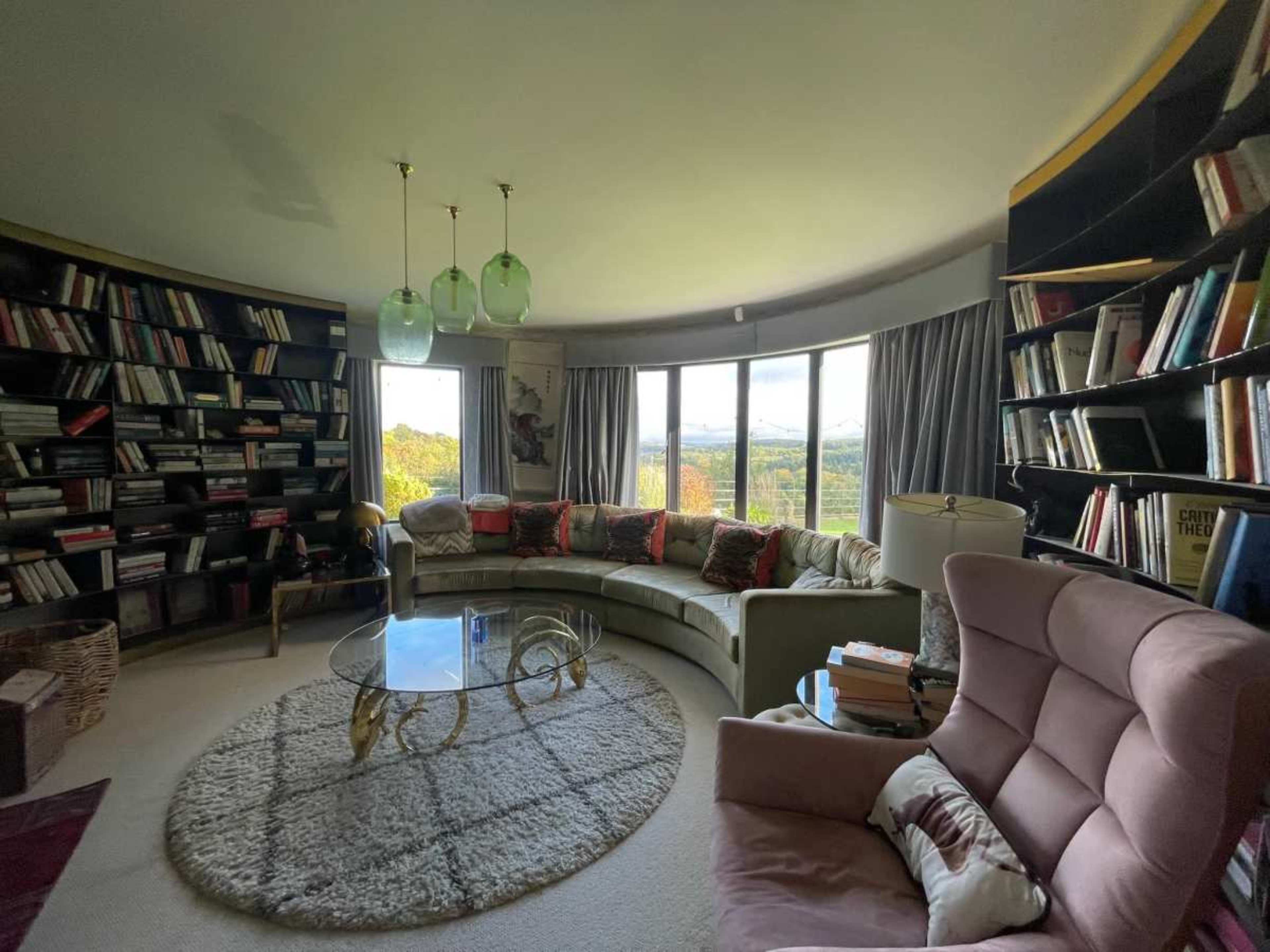 The image shows a cozy living room with a curved sectional sofa, a glass coffee table, and floor-to-ceiling bookshelves filled with books, overlooking a scenic view through large windows.
