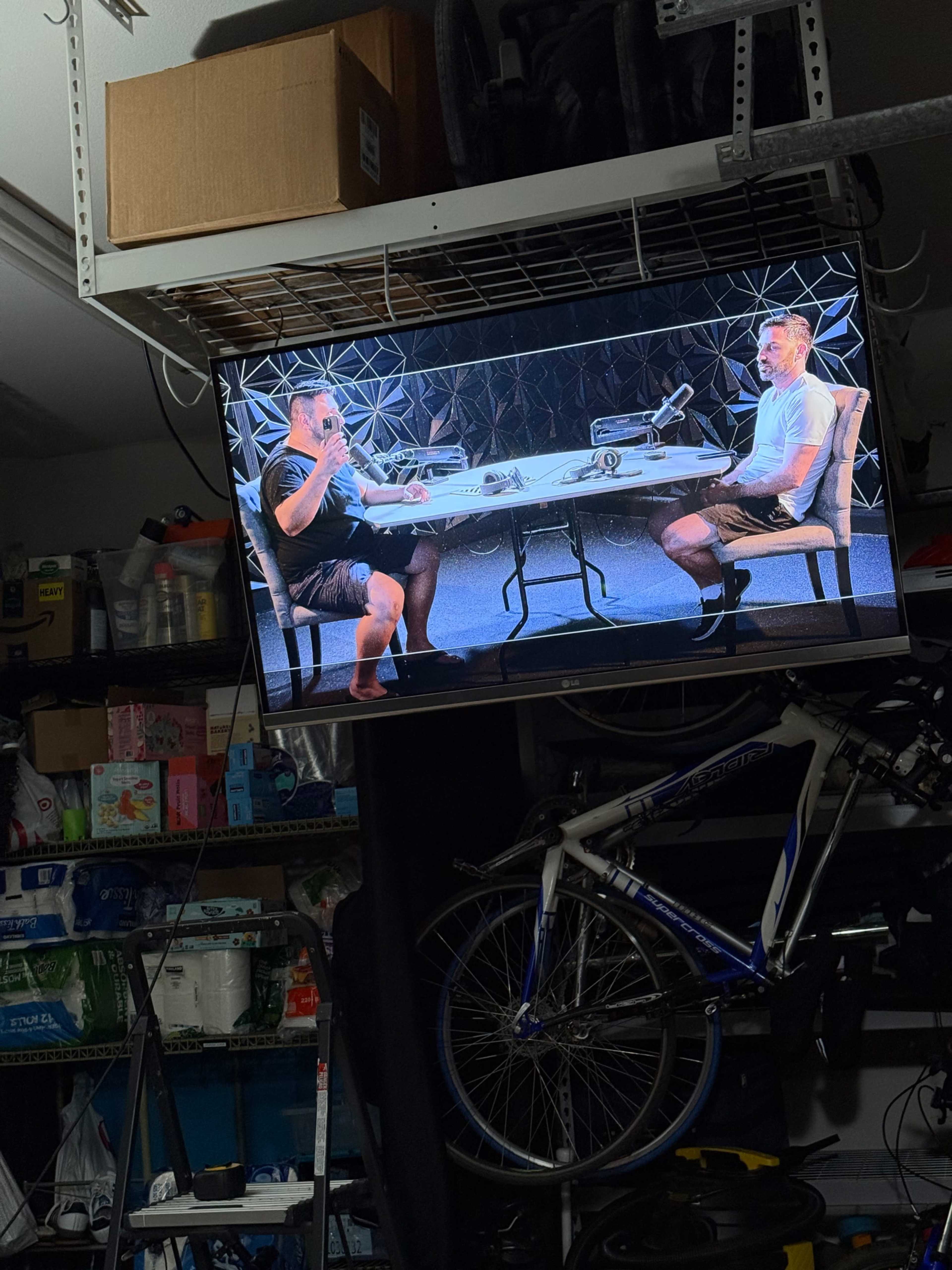 A television in a garage displays two people seated at a table with microphones, engaged in conversation.