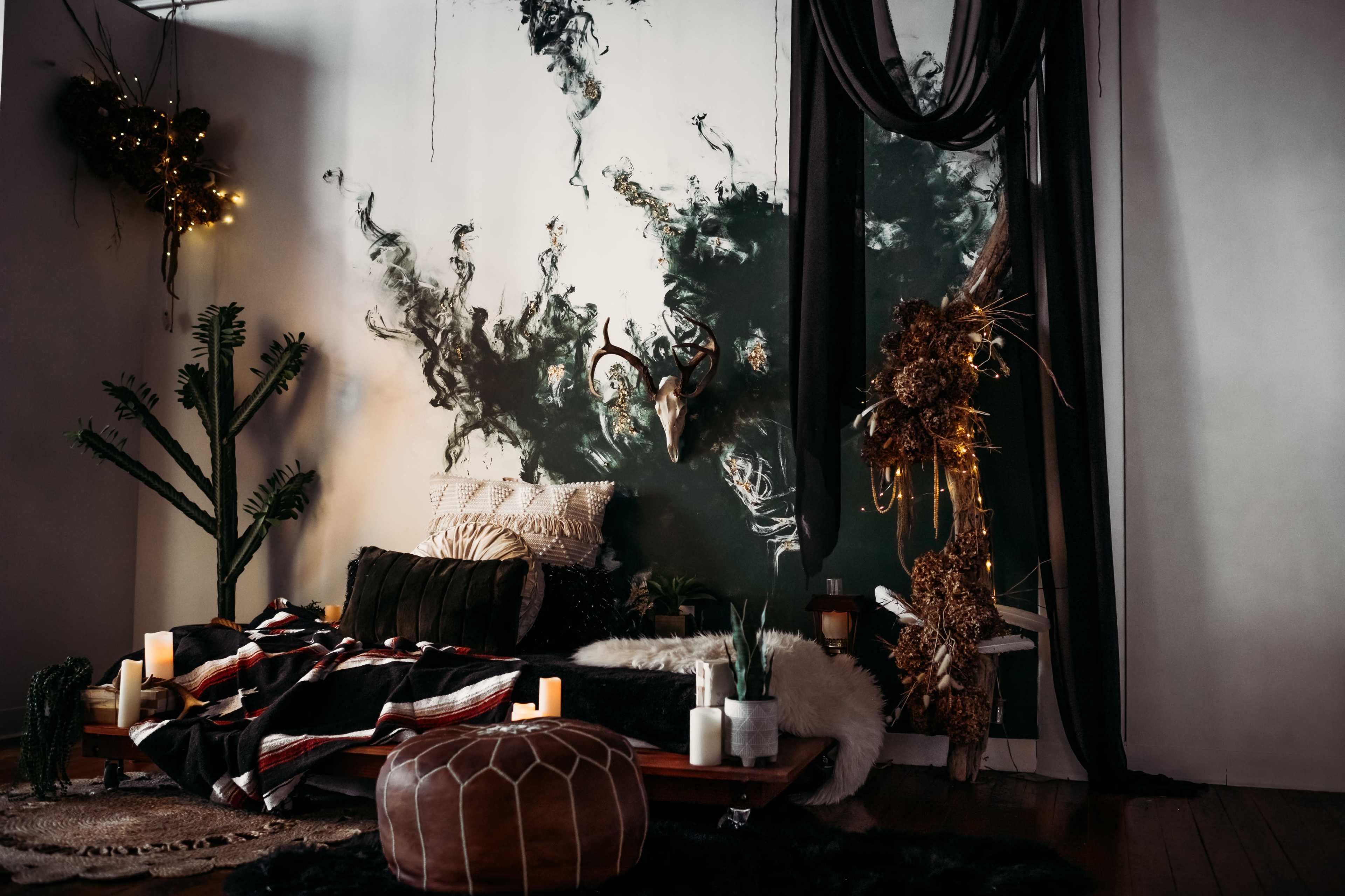 A cozy indoor space featuring a low wooden bed with a dark blanket, adorned with candles and decorative elements, against a wall displaying an abstract mural and surrounded by plants and natural decorations.