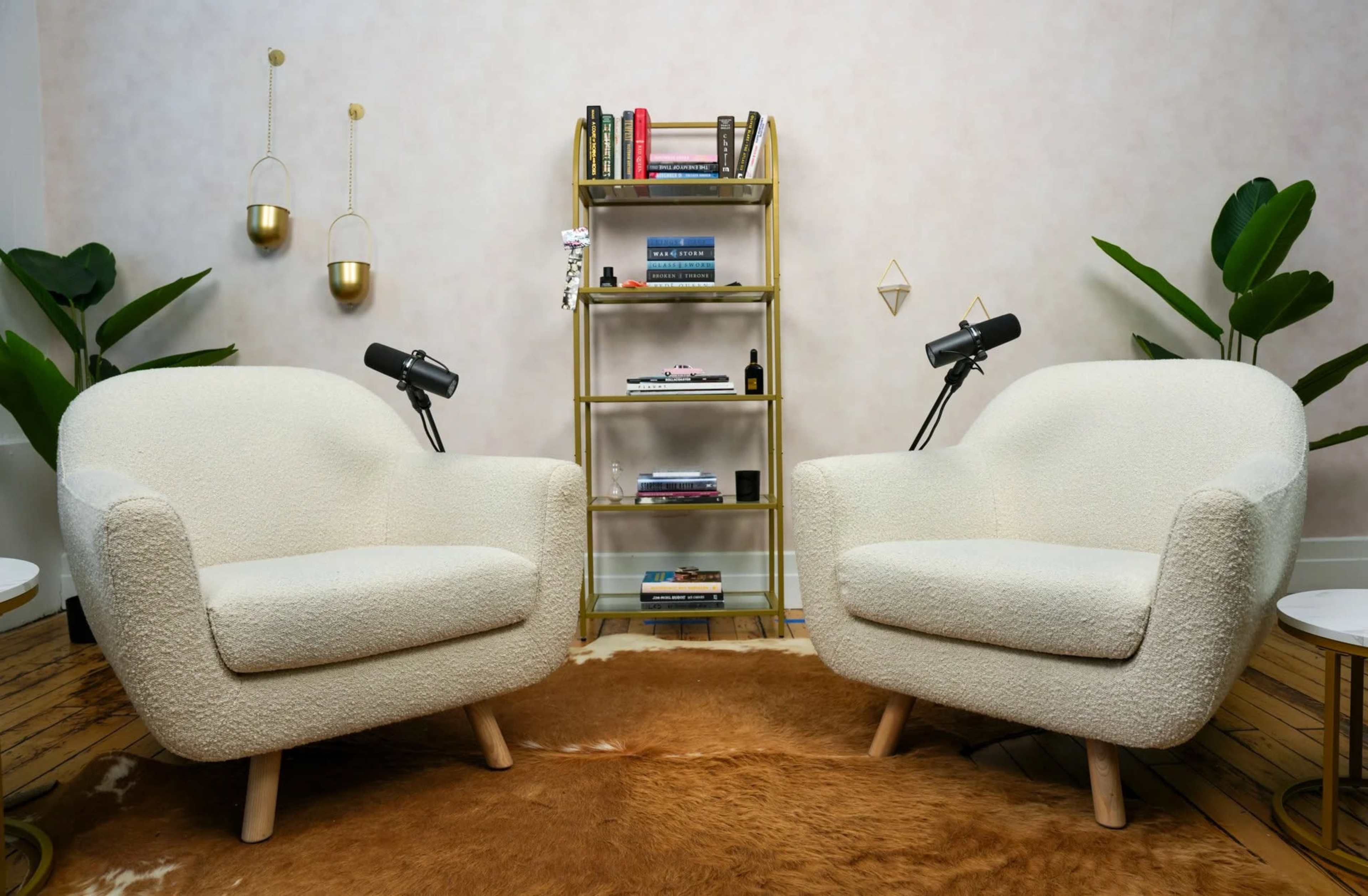 The image features two white, plush chairs facing each other over a brown rug, with a gold bookshelf and decorative plants in the background.