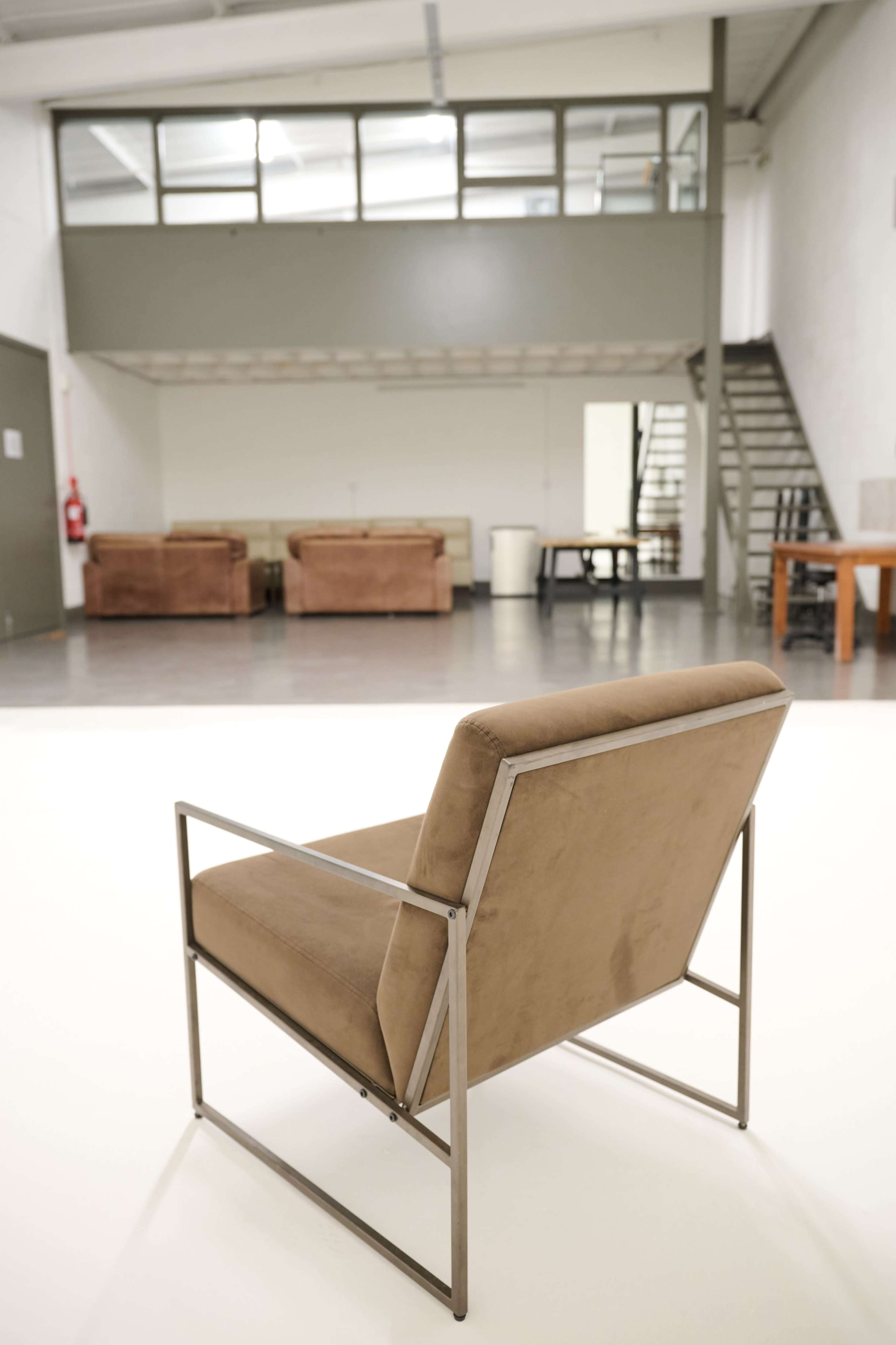 A single modern chair with a metal frame and fabric upholstery is positioned in a spacious, minimalist room featuring a staircase and seating area in the background.