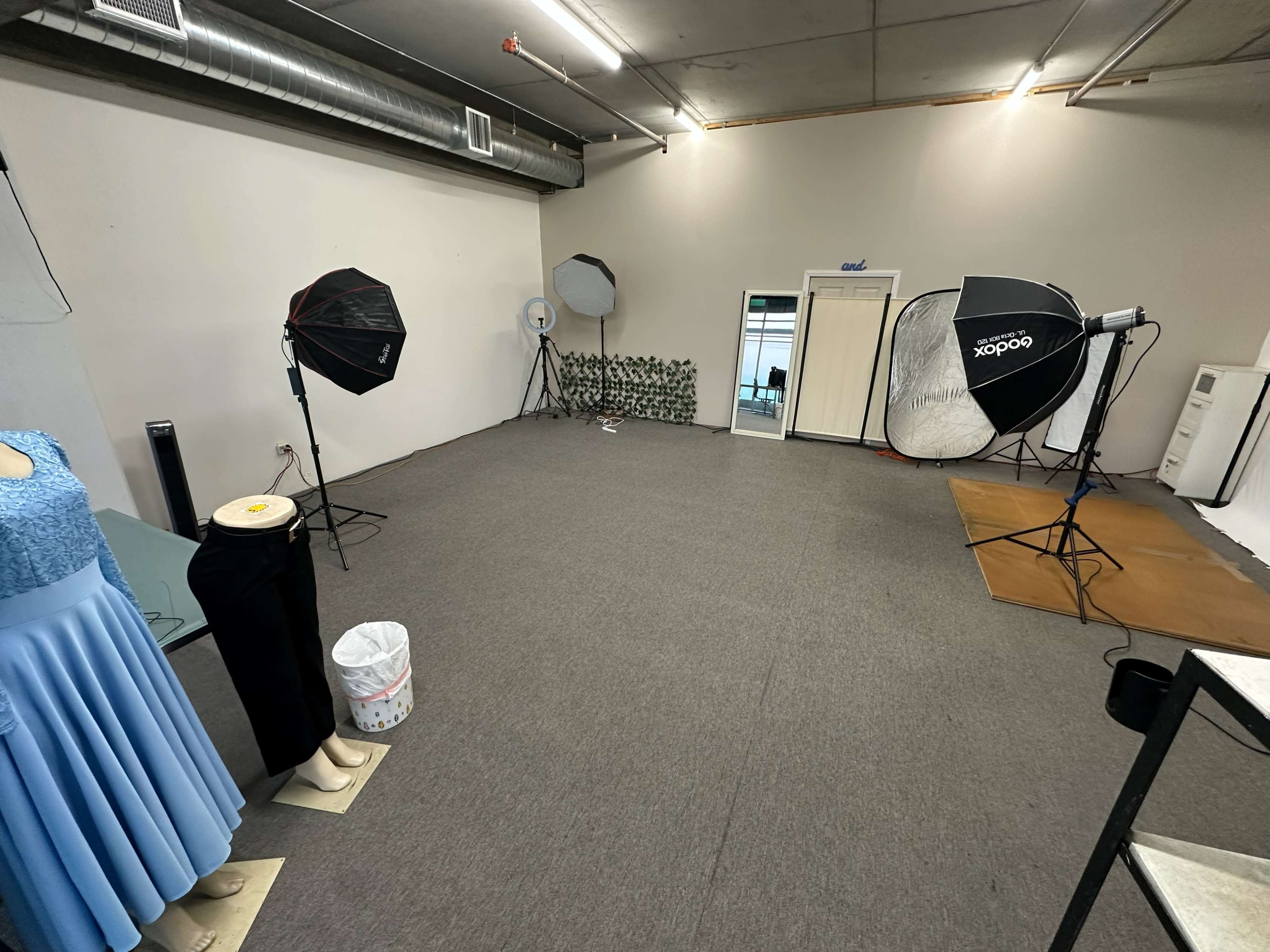 The image shows a spacious, empty studio with photography equipment, including softbox lights and a mirror, set against plain walls and carpeting.