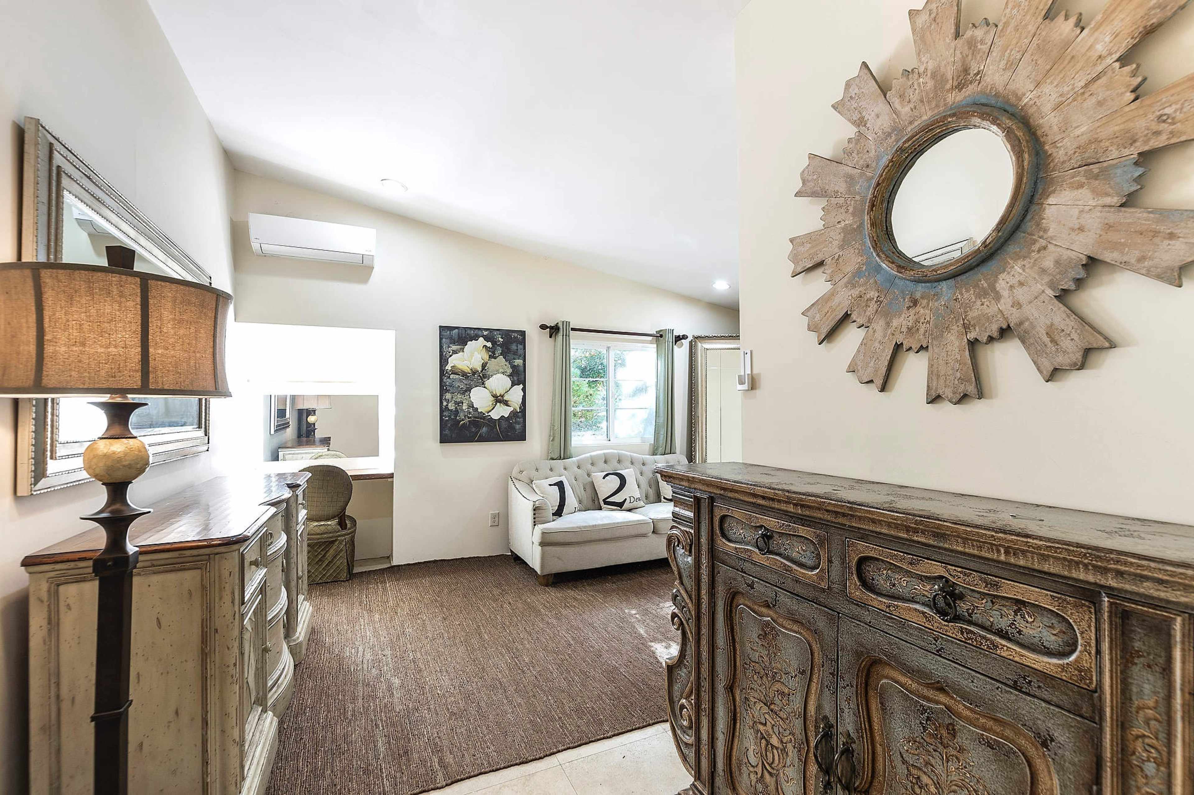 The image shows a well-lit living space featuring a decorative mirror, a wooden dresser, a couch with cushions, and a large wall art piece.