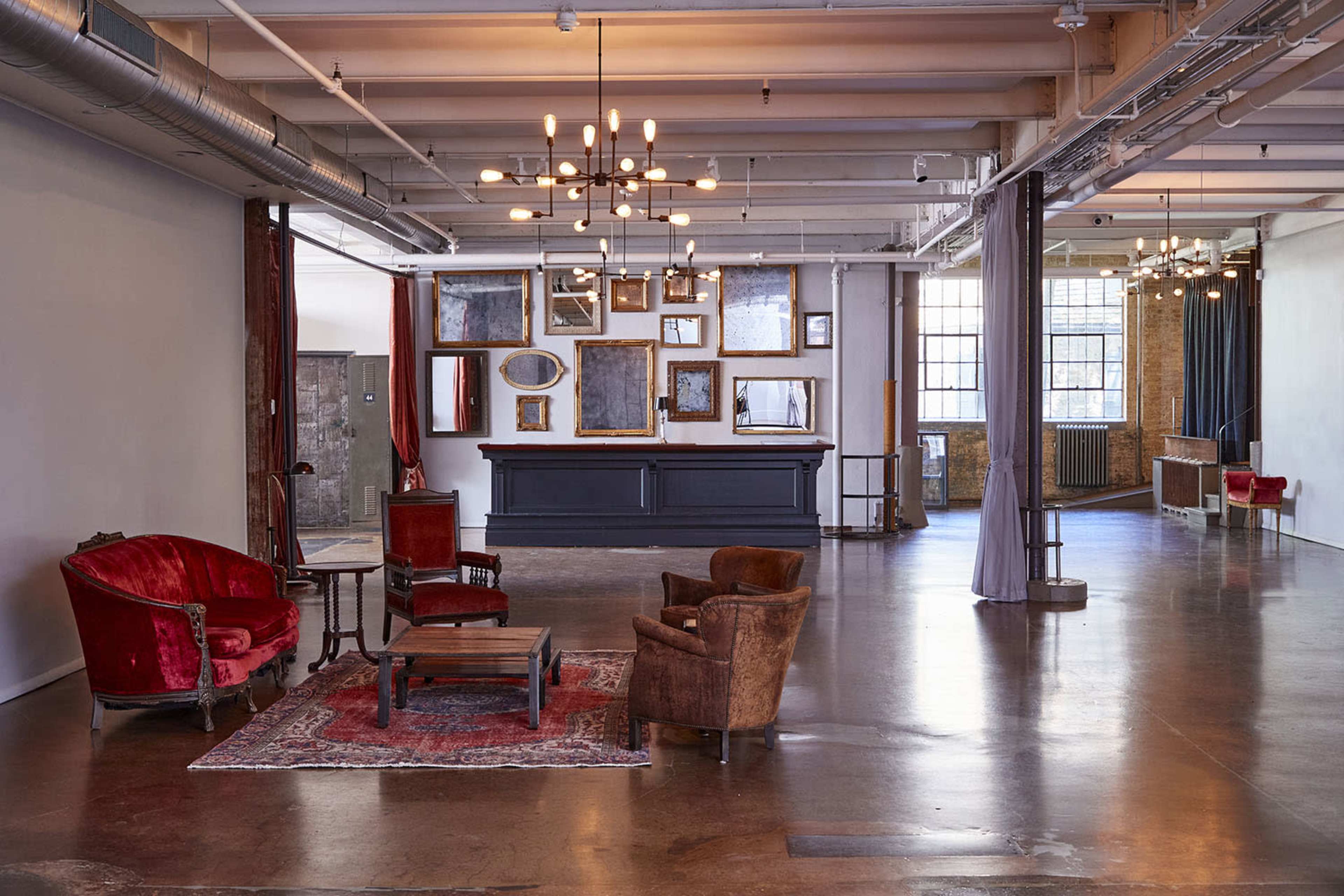 The image shows a spacious, well-lit industrial loft with a wooden floor, vintage furnishings, and a gallery-like wall adorned with various frames.