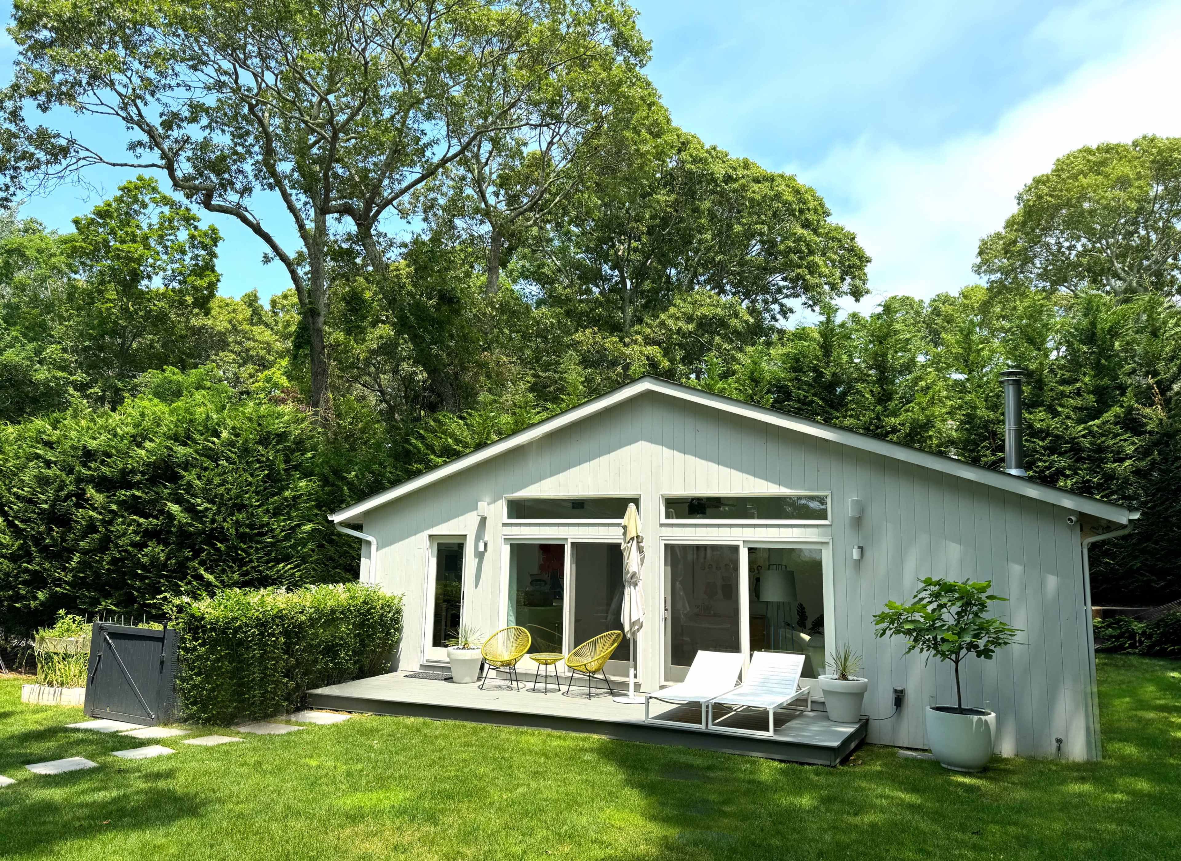 A modern single-story house features large windows and a deck with lounge chairs, surrounded by lush greenery.
