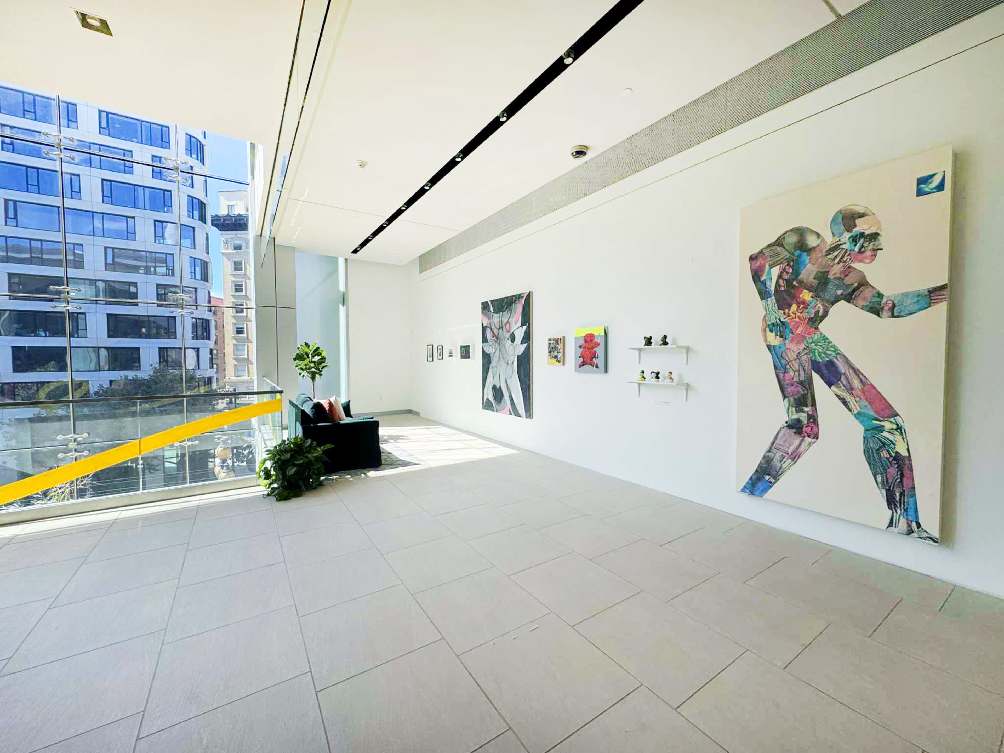 The image shows a modern art gallery with large windows, featuring colorful artwork on the walls and a small seating area.