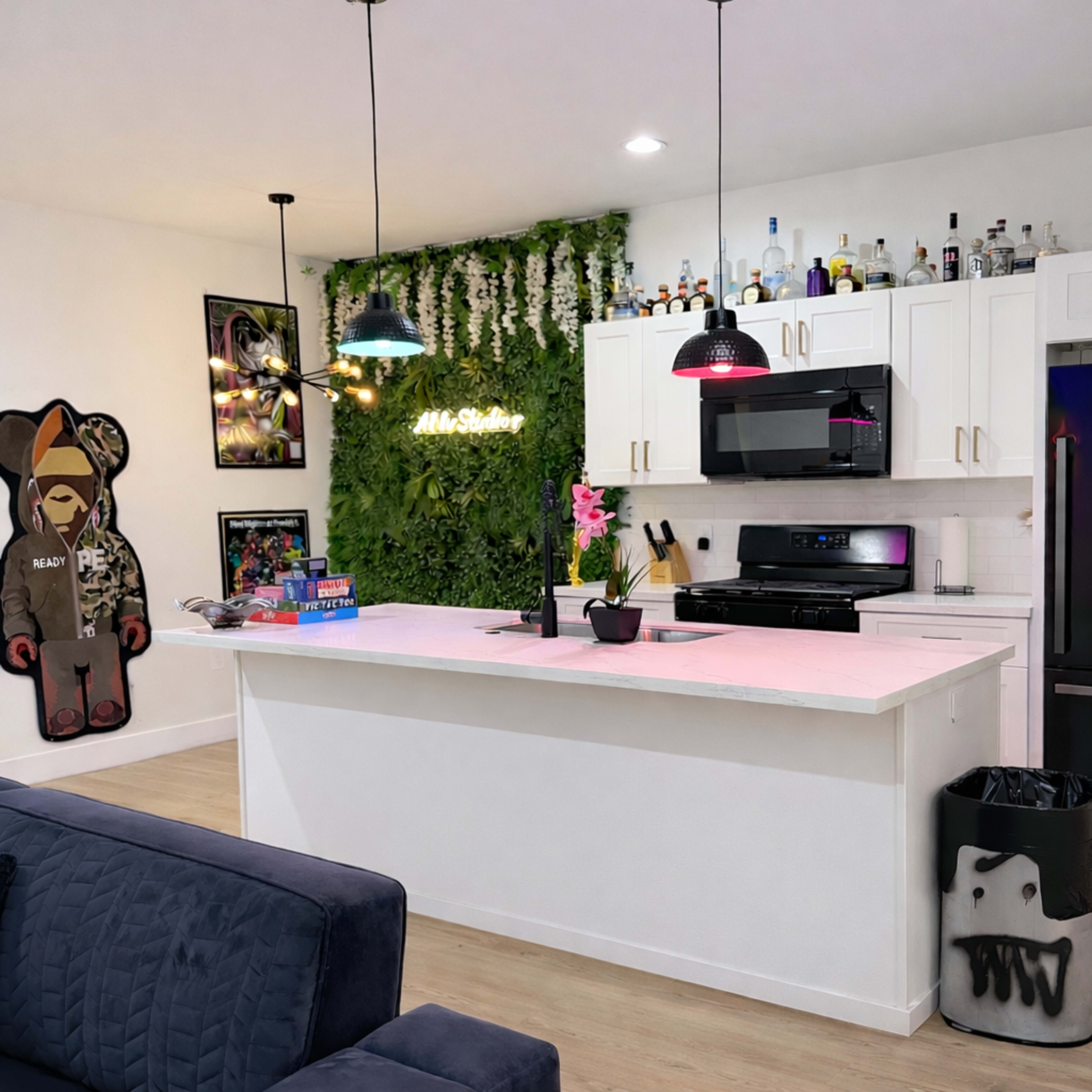 The image shows a modern kitchen with white cabinets, a large island with seating, and a green wall of plants, complemented by decorative bottles and artwork on the walls.