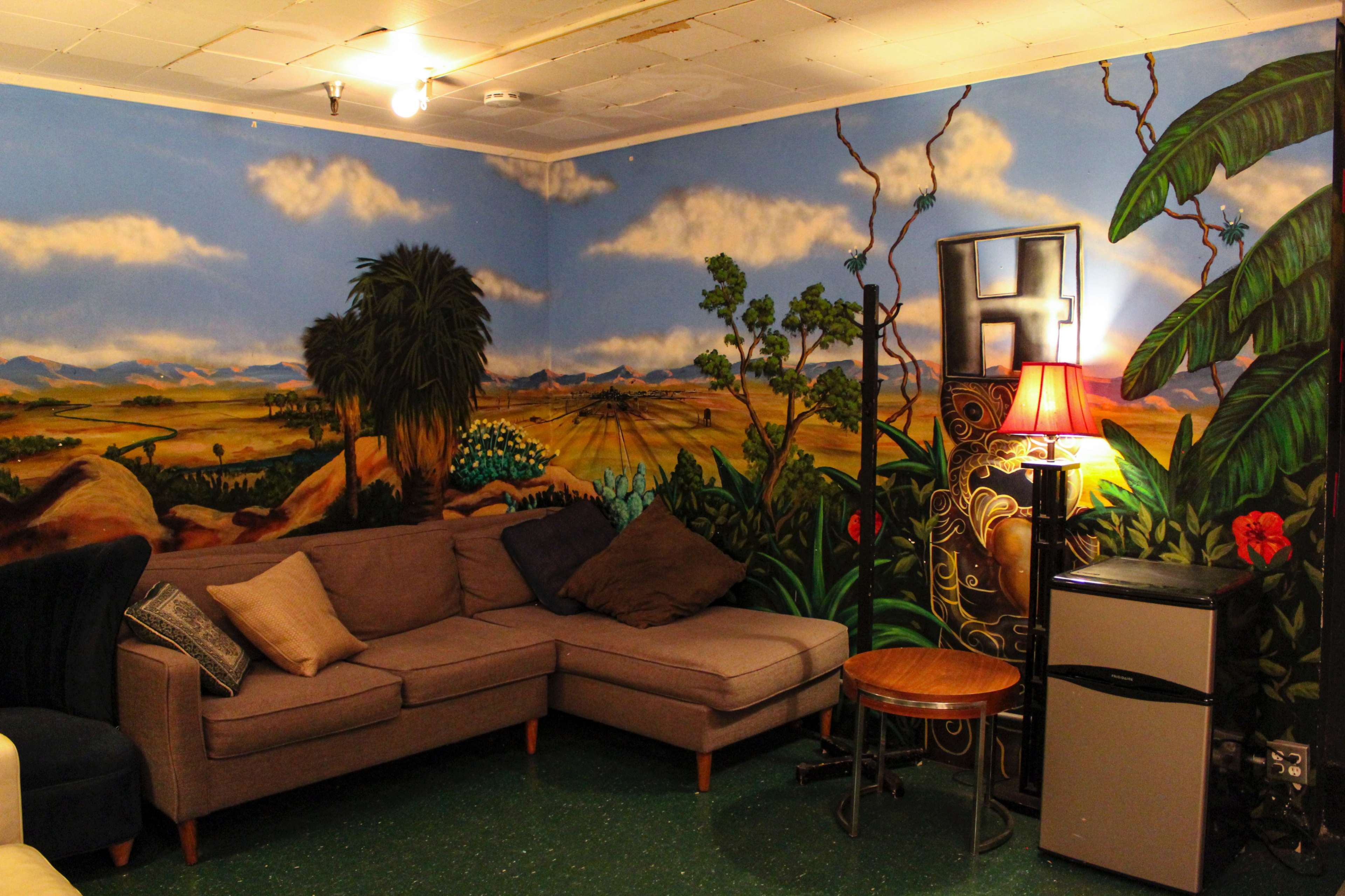 The image depicts a room with a large mural of a landscape, featuring mountains and greenery, alongside a sectional sofa, a small table, a lamp, and a mini refrigerator.