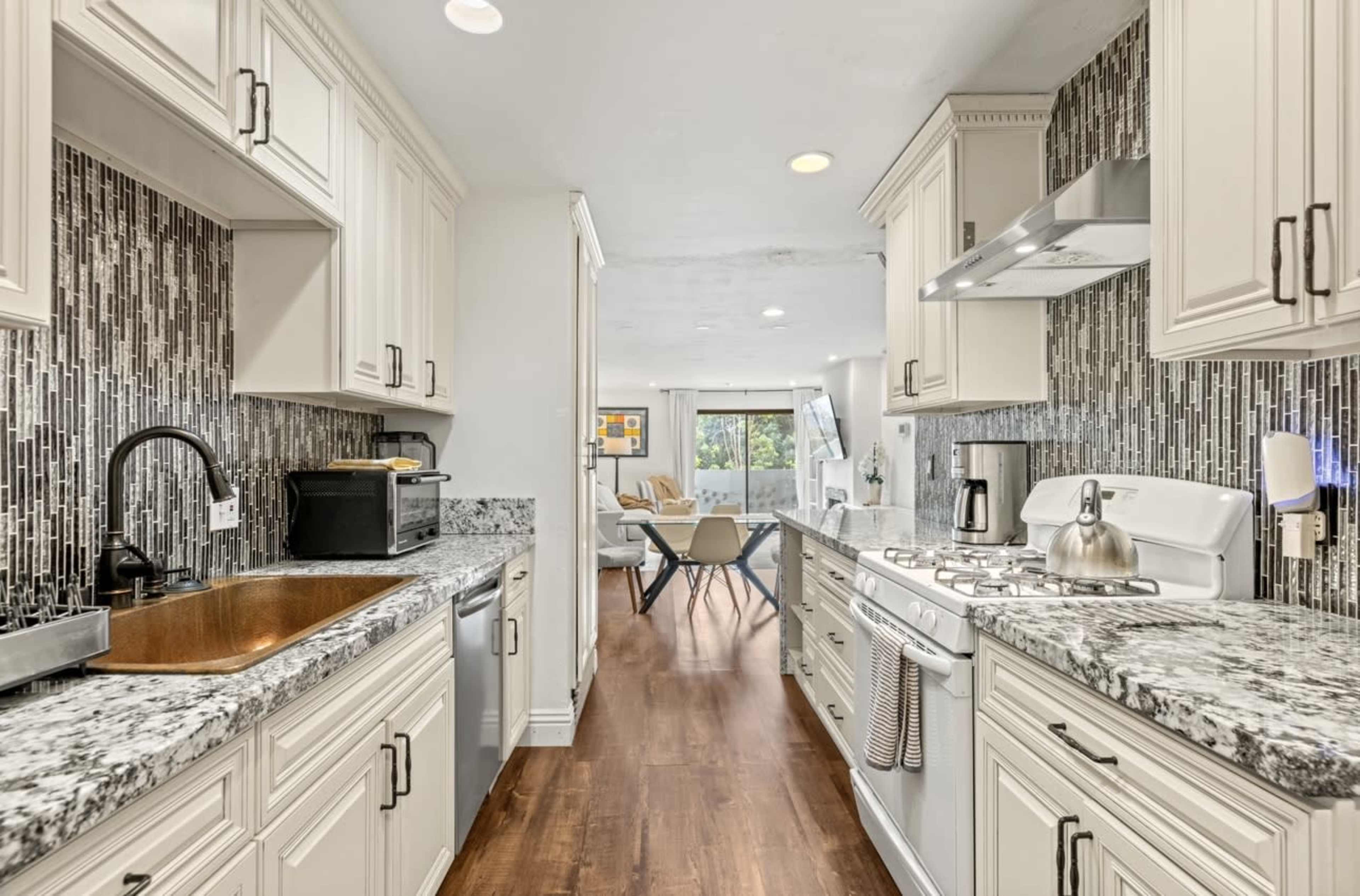The image shows a modern kitchen with granite countertops, white cabinetry, and a tile backsplash, leading to a living area with a large window and contemporary furniture.