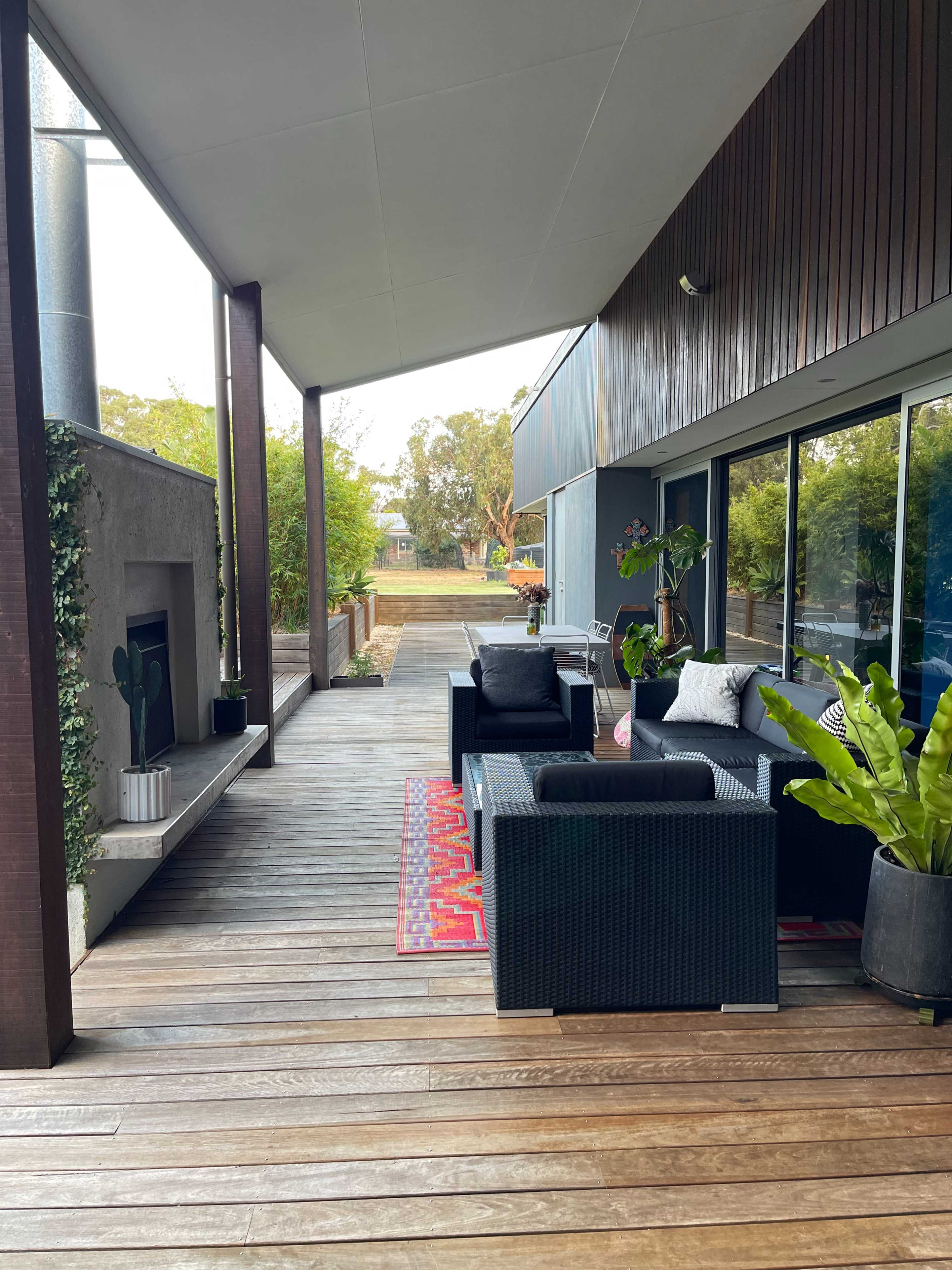 The image shows a modern outdoor living area with black wicker furniture arranged on a wooden deck, featuring a fireplace and greenery in the background.