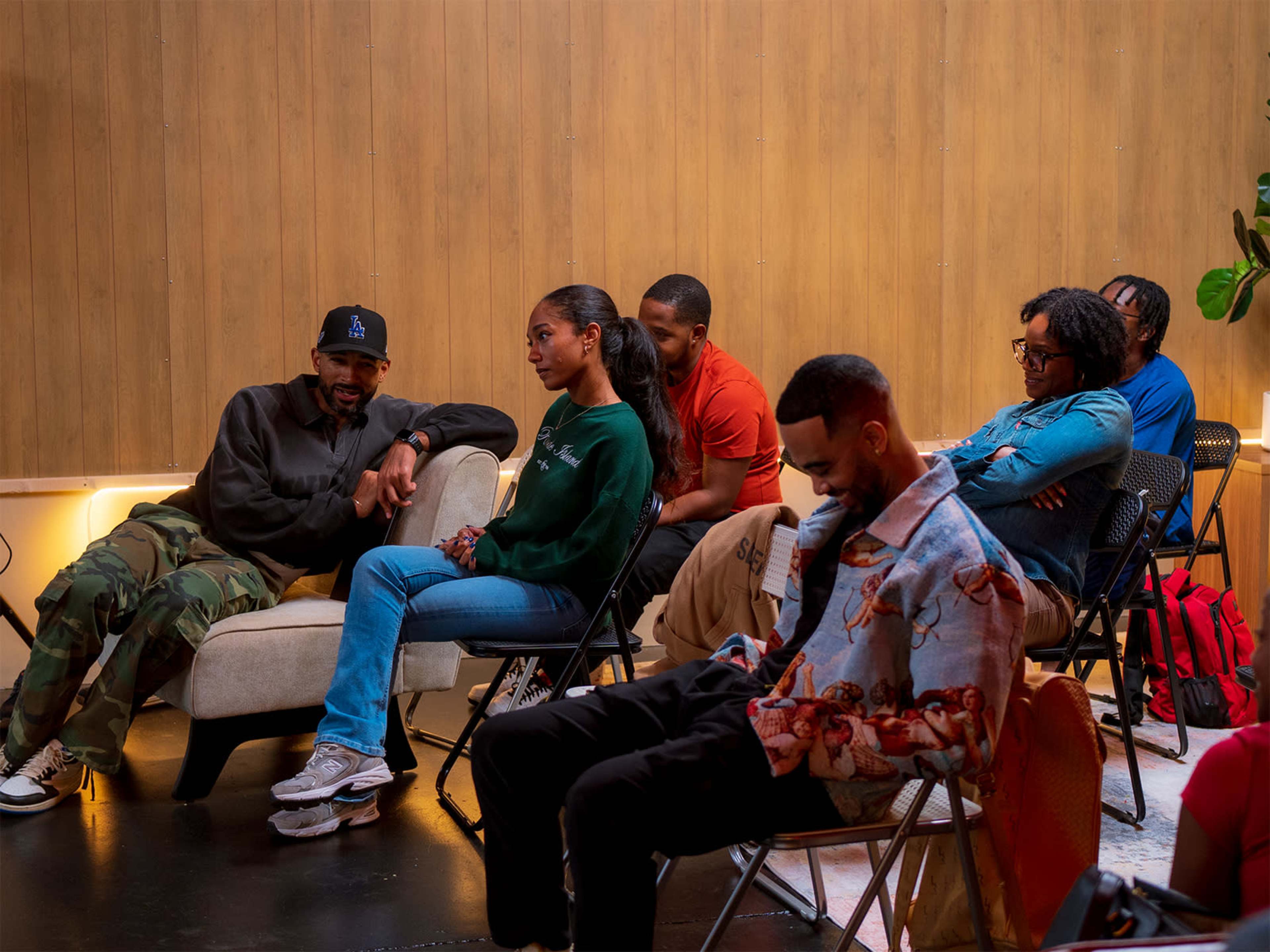 A group of six people sit in folding chairs in a small, well-lit venue with wooden walls, while two are engaged in conversation and the others appear to be listening or using their phones.