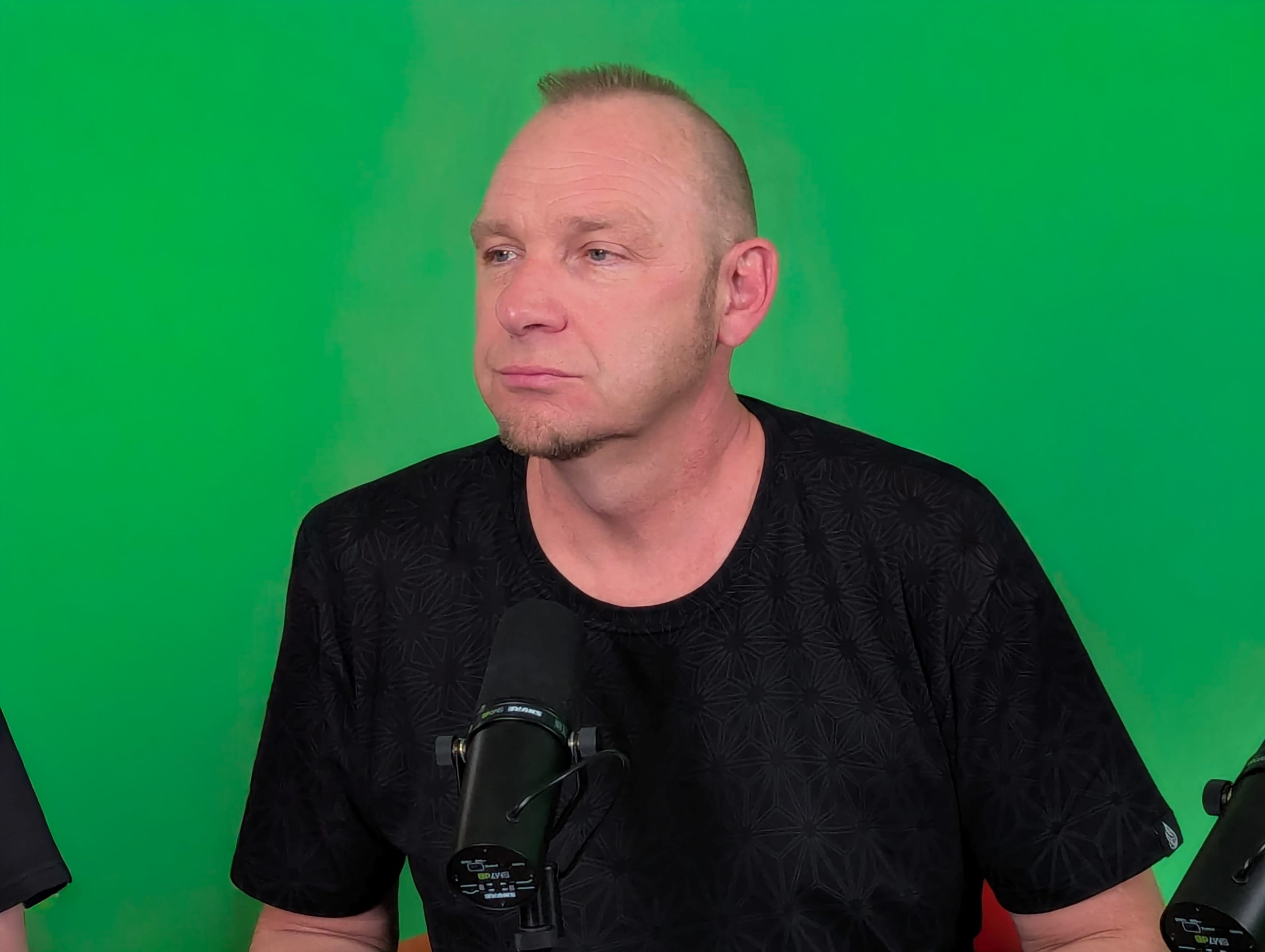 A man with short hair and a black patterned shirt sits in front of a green backdrop with microphones positioned nearby.
