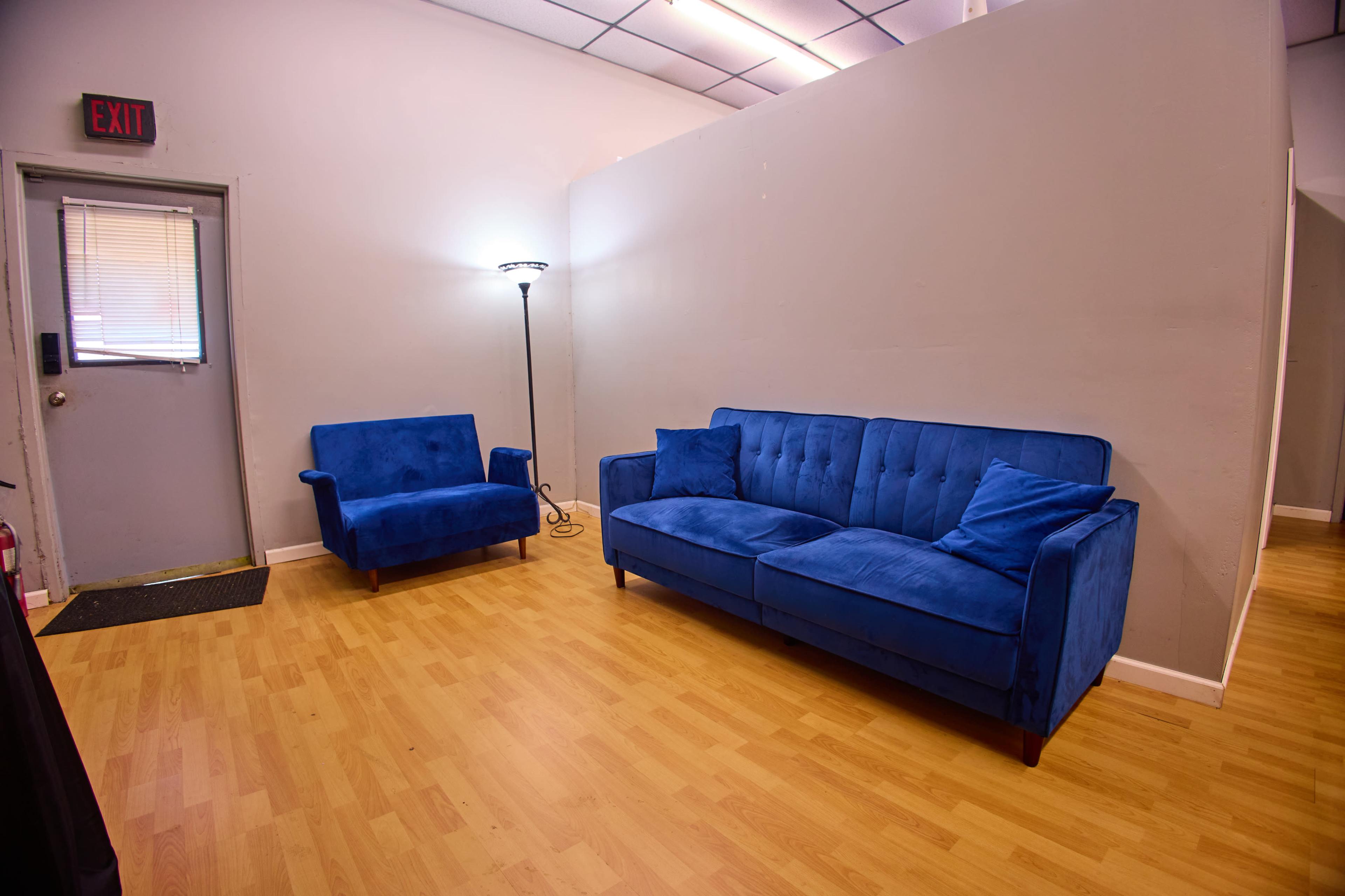 A blue couch and a matching chair are positioned against a light gray wall in a room with a wooden floor and a single door.