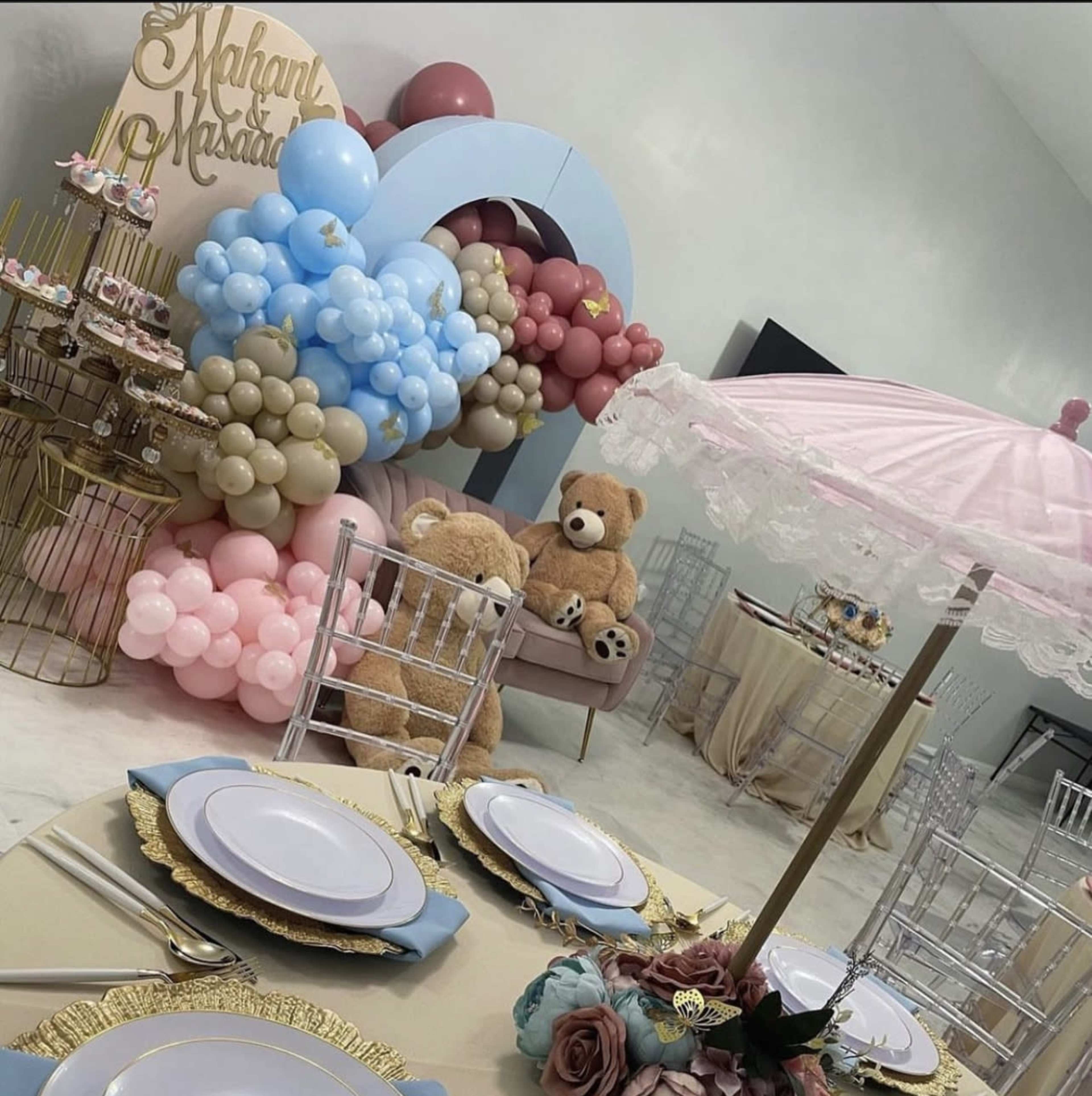 The scene features a decorated party space with pastel-colored balloons, a dessert table with cupcakes, and two teddy bears sitting on a plush seating area.