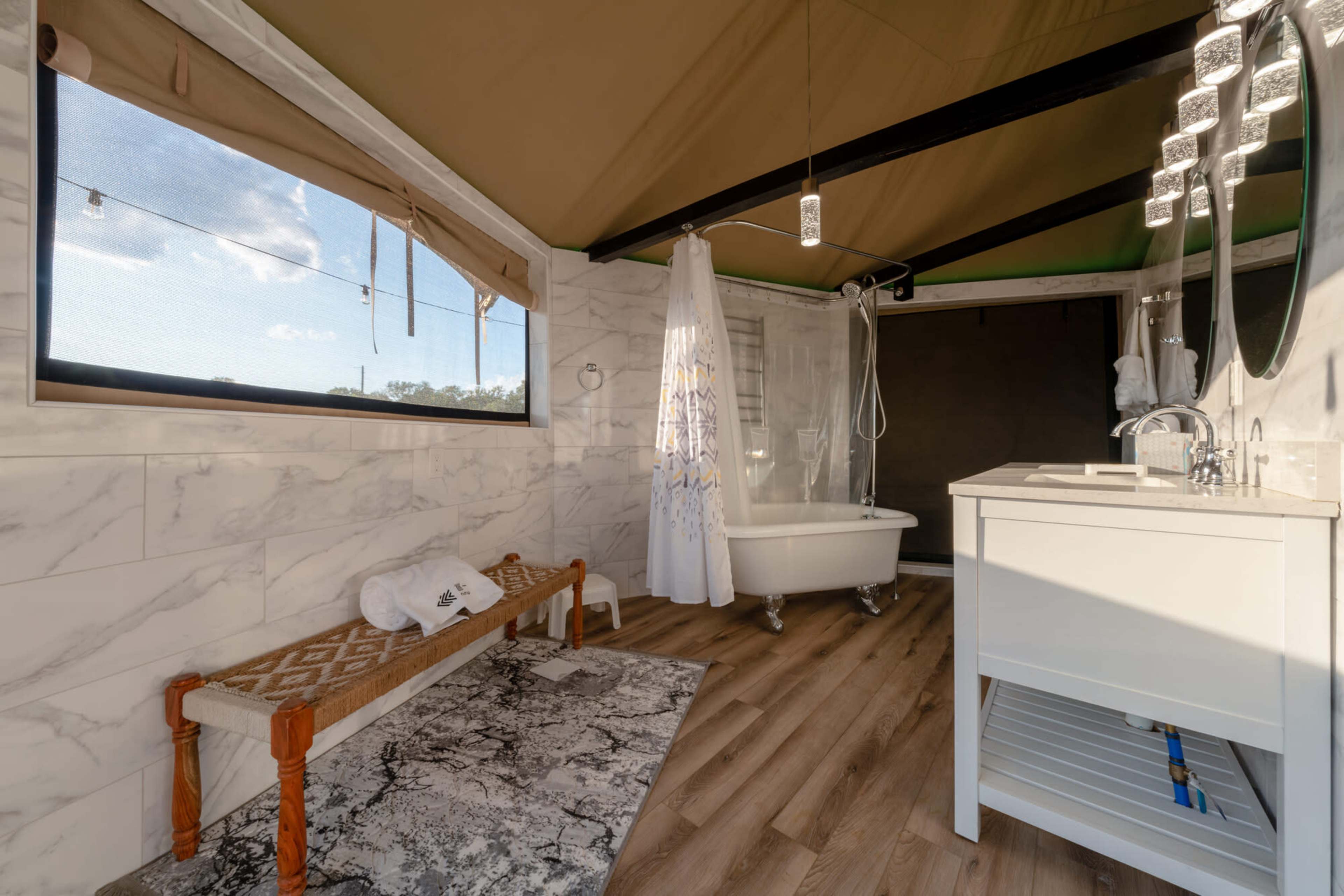 The image shows a modern bathroom with a freestanding bathtub, a white vanity, and a shower enclosed by a curtain, set against a backdrop of marble-style walls and wooden flooring.
