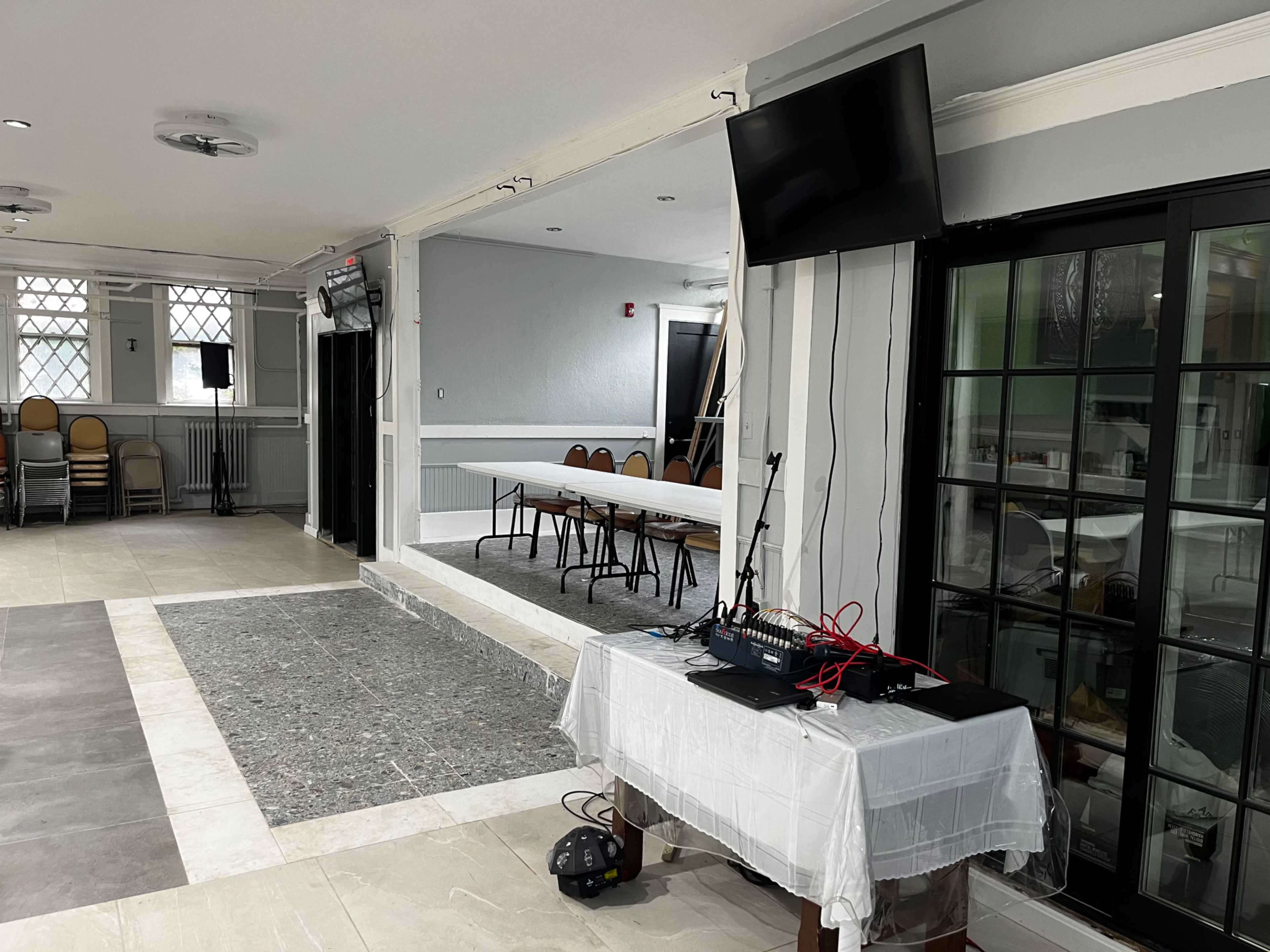 A spacious interior room features a setup for an event with a table for equipment and a large table in the background, flanked by chairs.