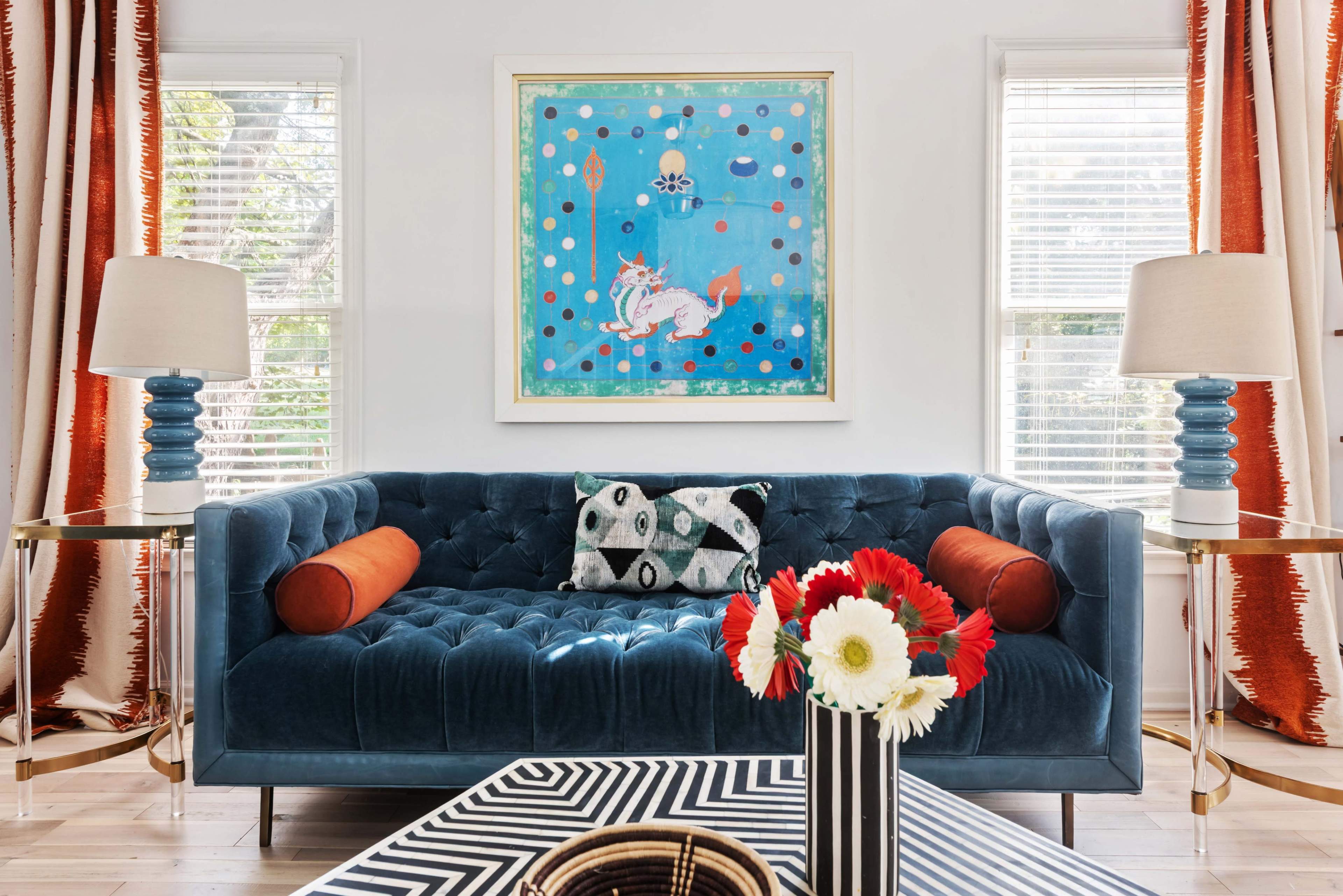The image shows a modern living room featuring a blue tufted sofa with orange and patterned throw pillows, flanked by two lamps on side tables, and adorned with a colorful painting above the sofa and a striped coffee table in the foreground.