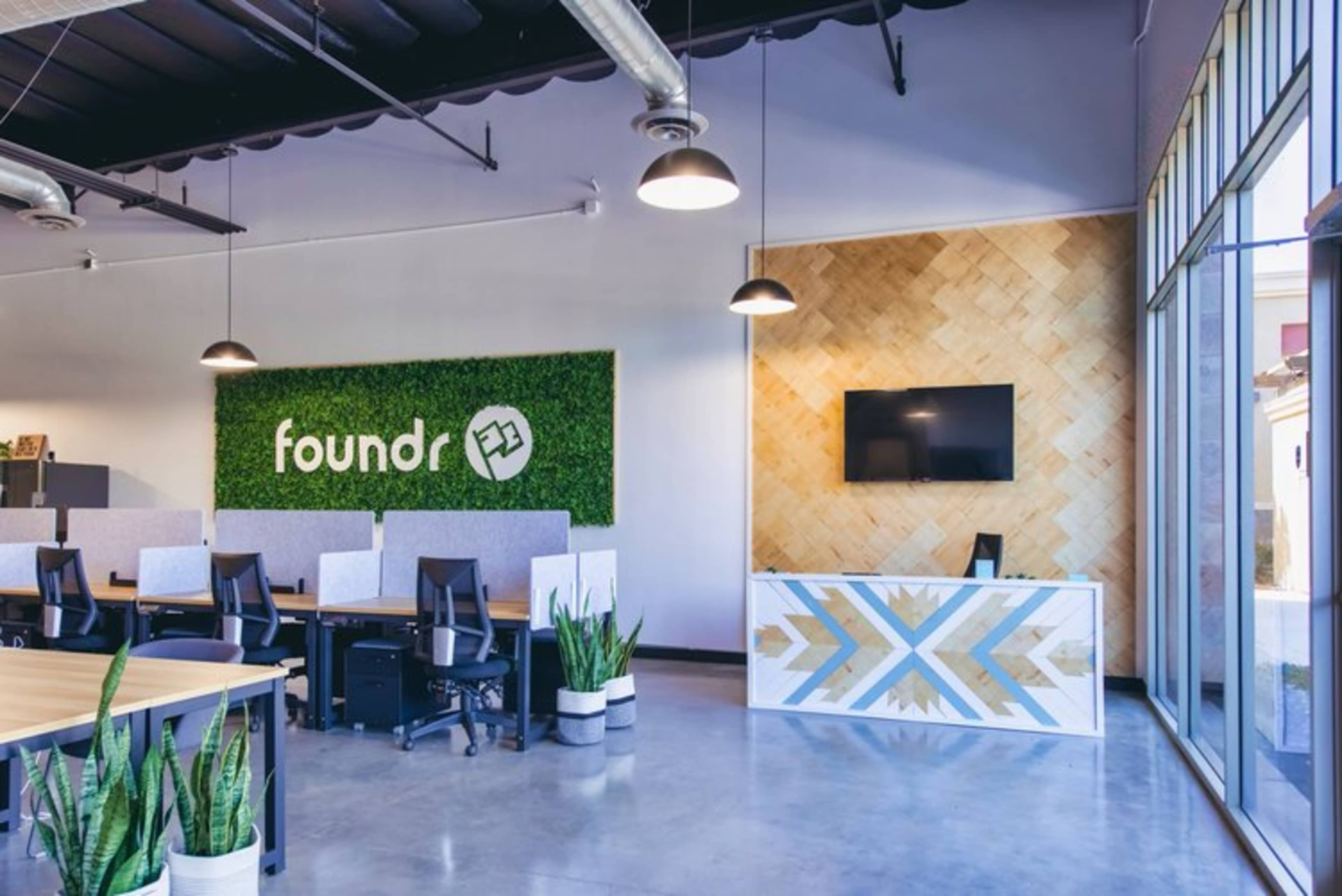 A modern office interior with workspace desks, a reception area with a decorative wall, and greenery elements.
