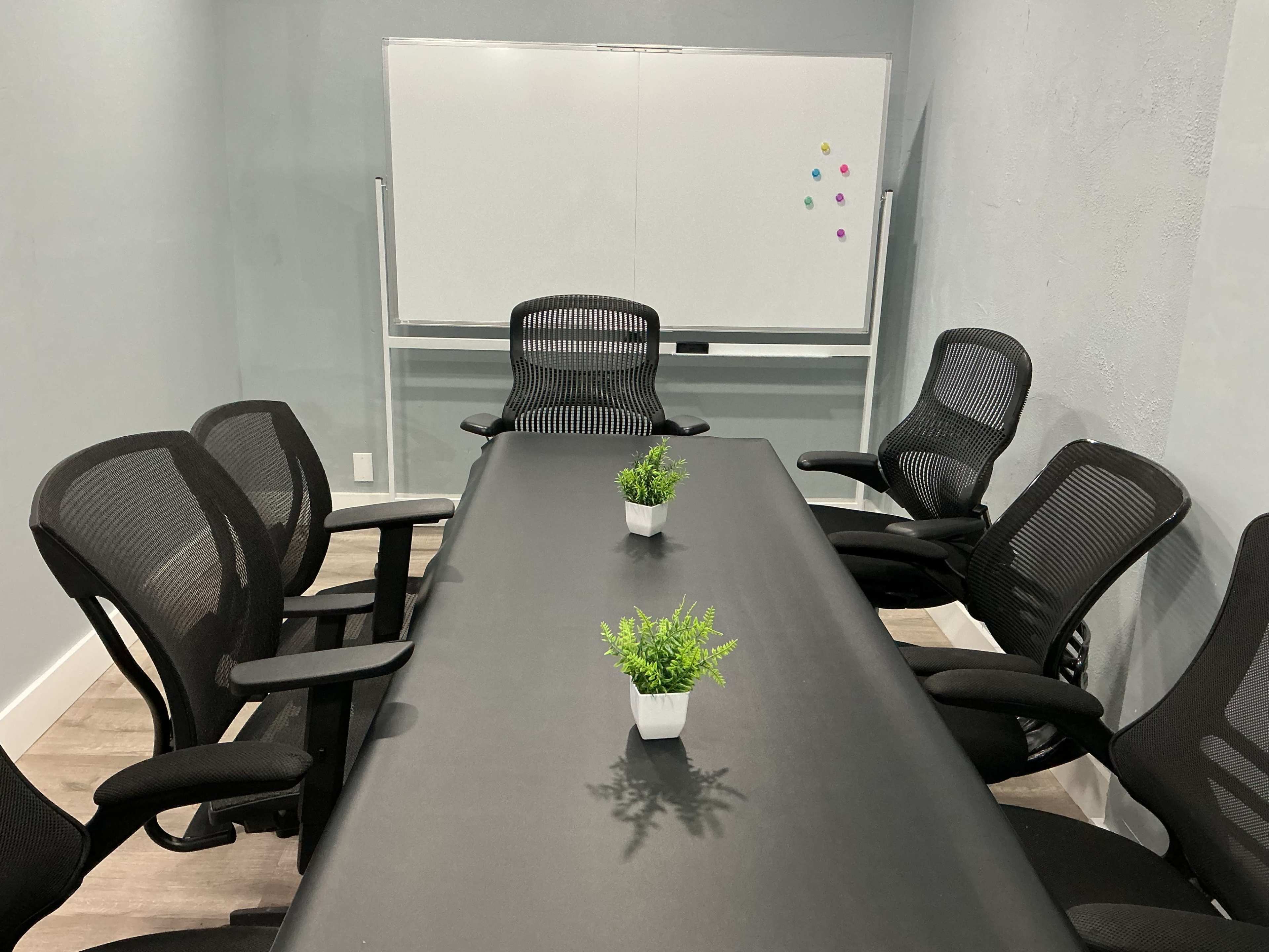 A conference room features a long black table surrounded by ergonomic office chairs and a whiteboard on the wall.