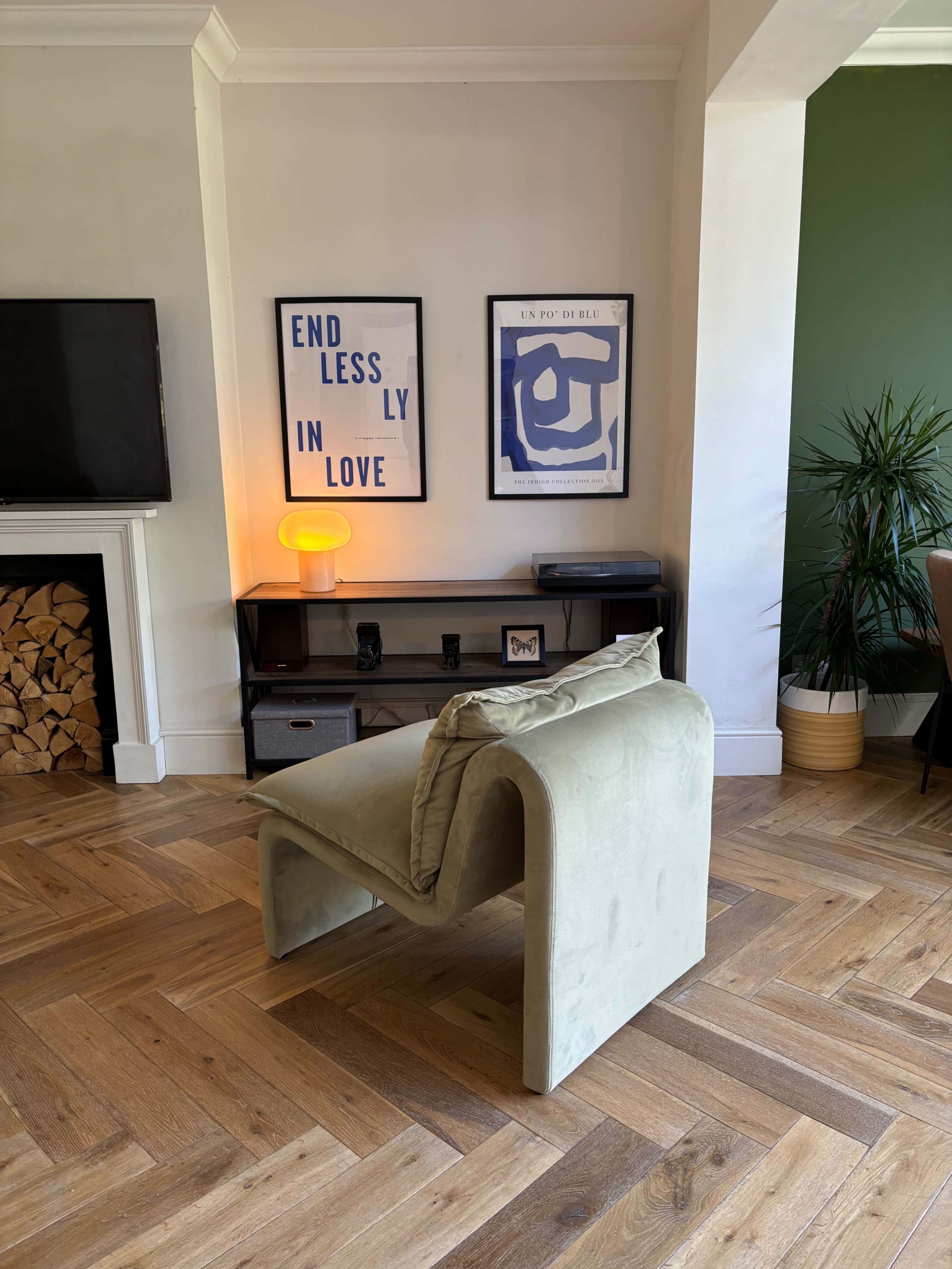 The image shows a modern living room with a curved green chair positioned near a television, a console table with decorative items, and framed artwork on the wall.