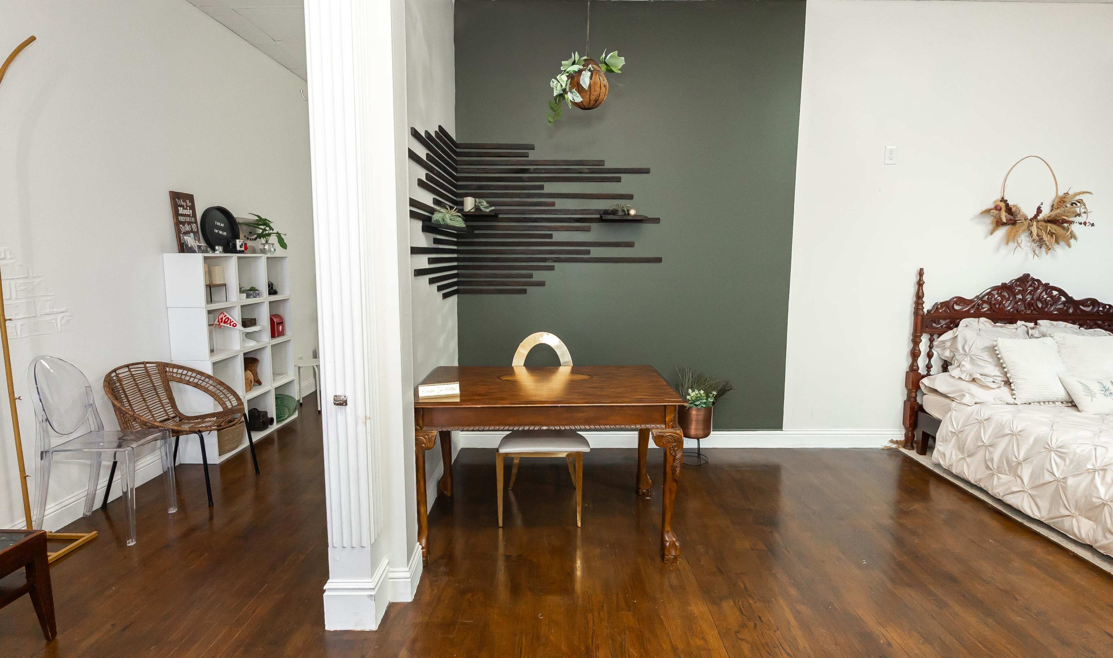 The image shows a cozy room featuring a wooden table with a decorative piece, a bed with light bedding, and a wall adorned with horizontal wooden slats and greenery.