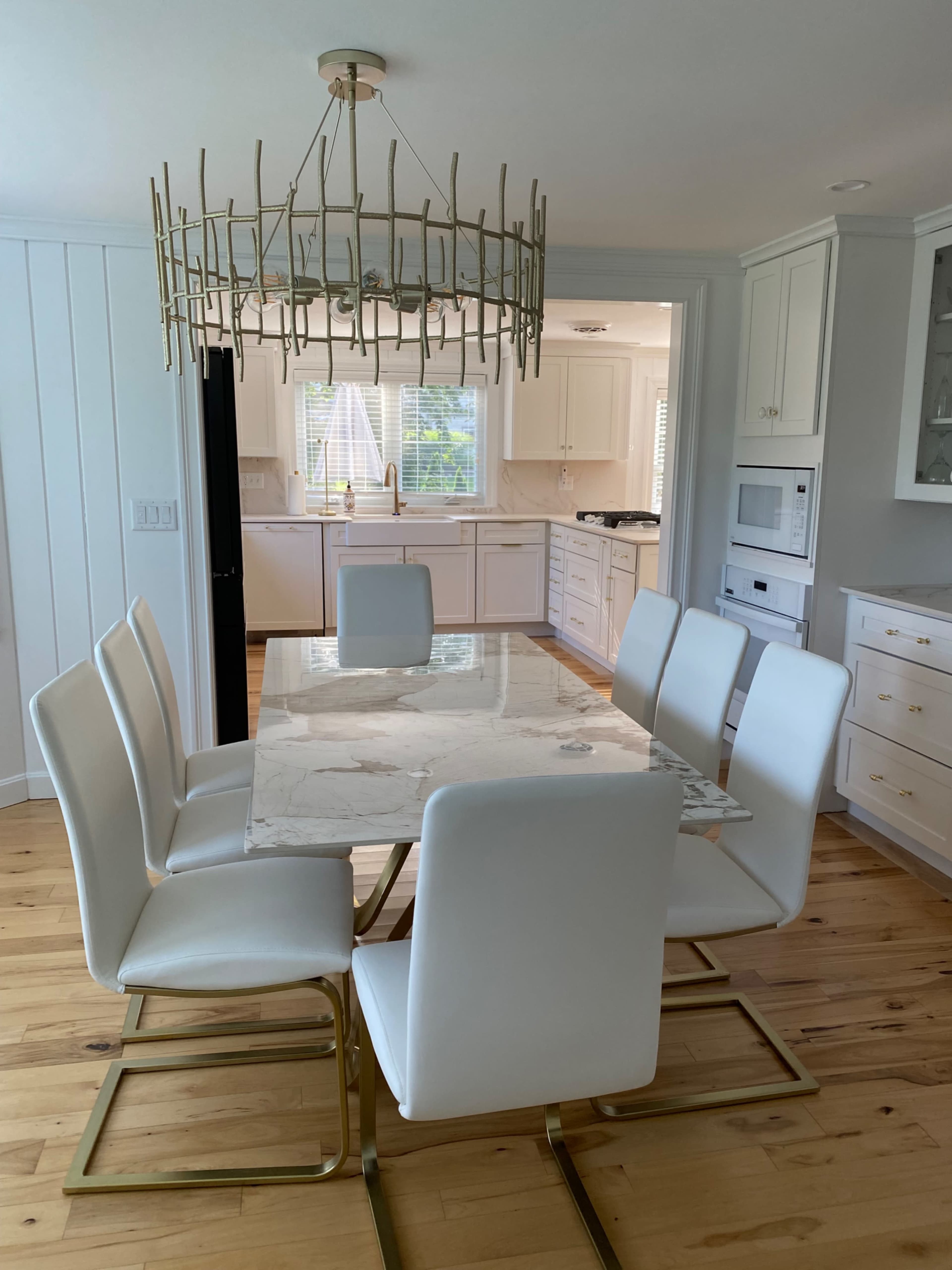 A modern dining area features a marble-topped table surrounded by white chairs, with a decorative chandelier overhead and a view of a kitchen in the background.