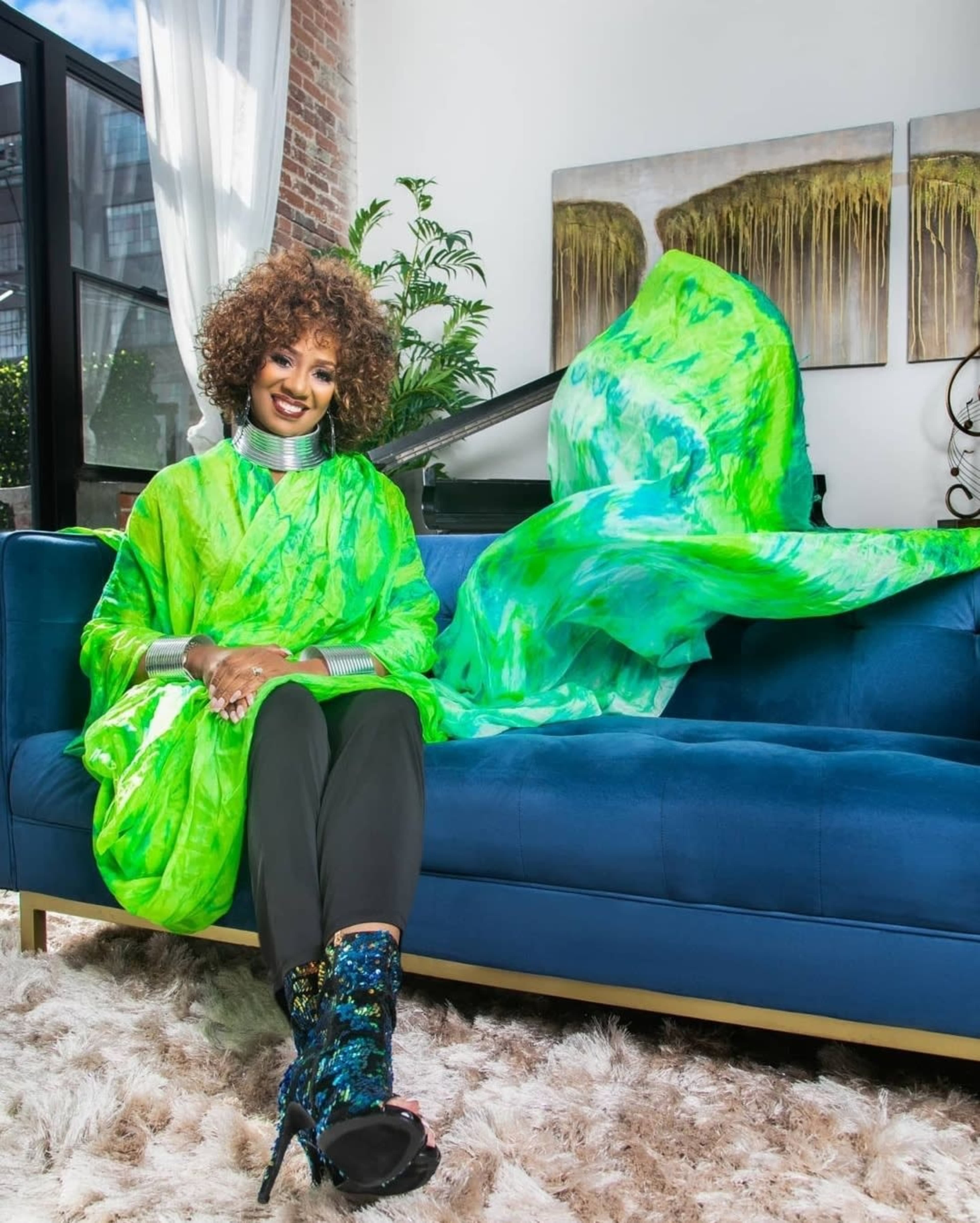 A person wearing a green outfit sits on a blue couch next to a flowing green fabric in a bright indoor space.