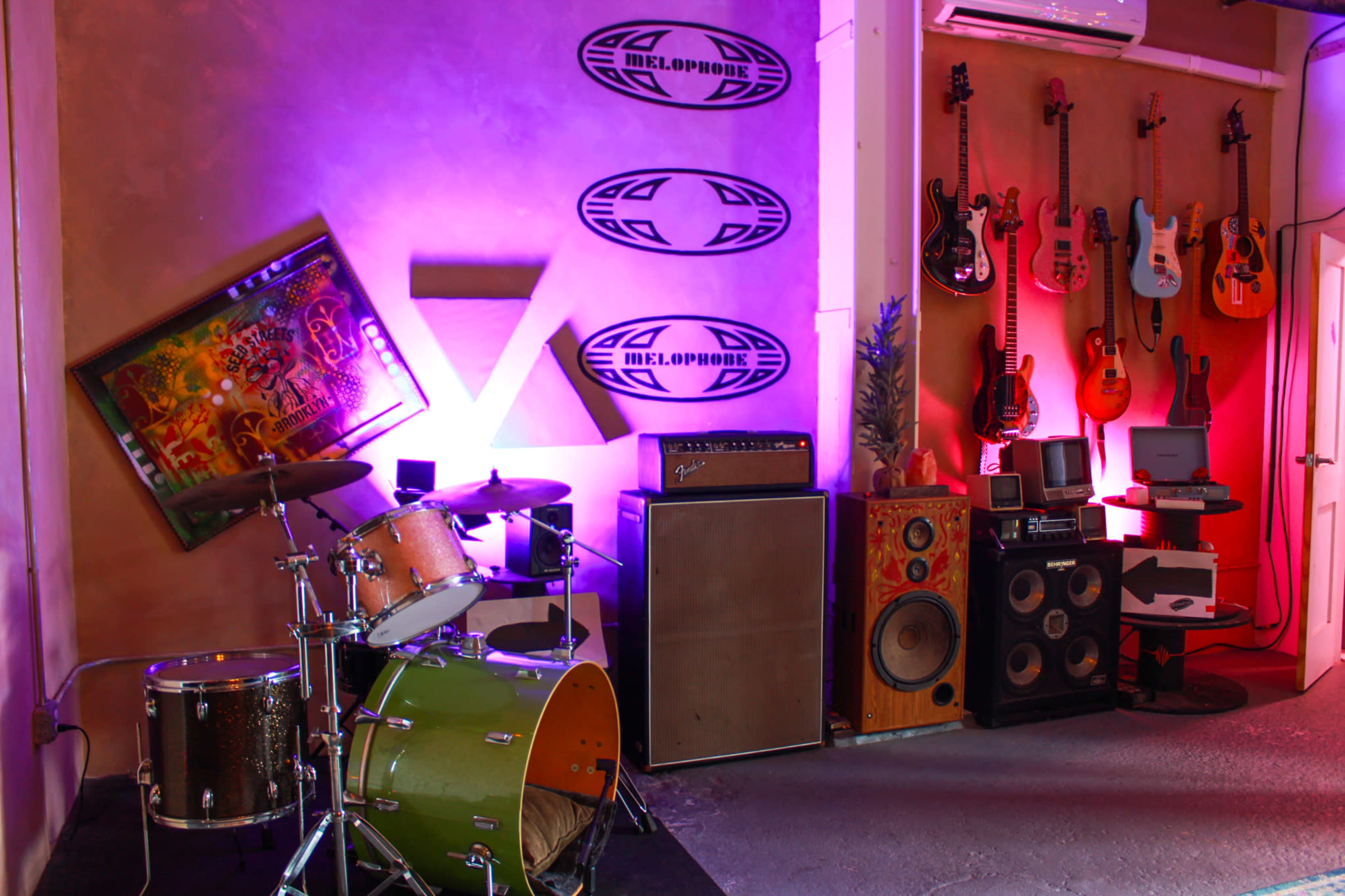 The image shows a music studio corner featuring a green drum set, multiple guitars hanging on the wall, an amplifier, and various audio equipment.