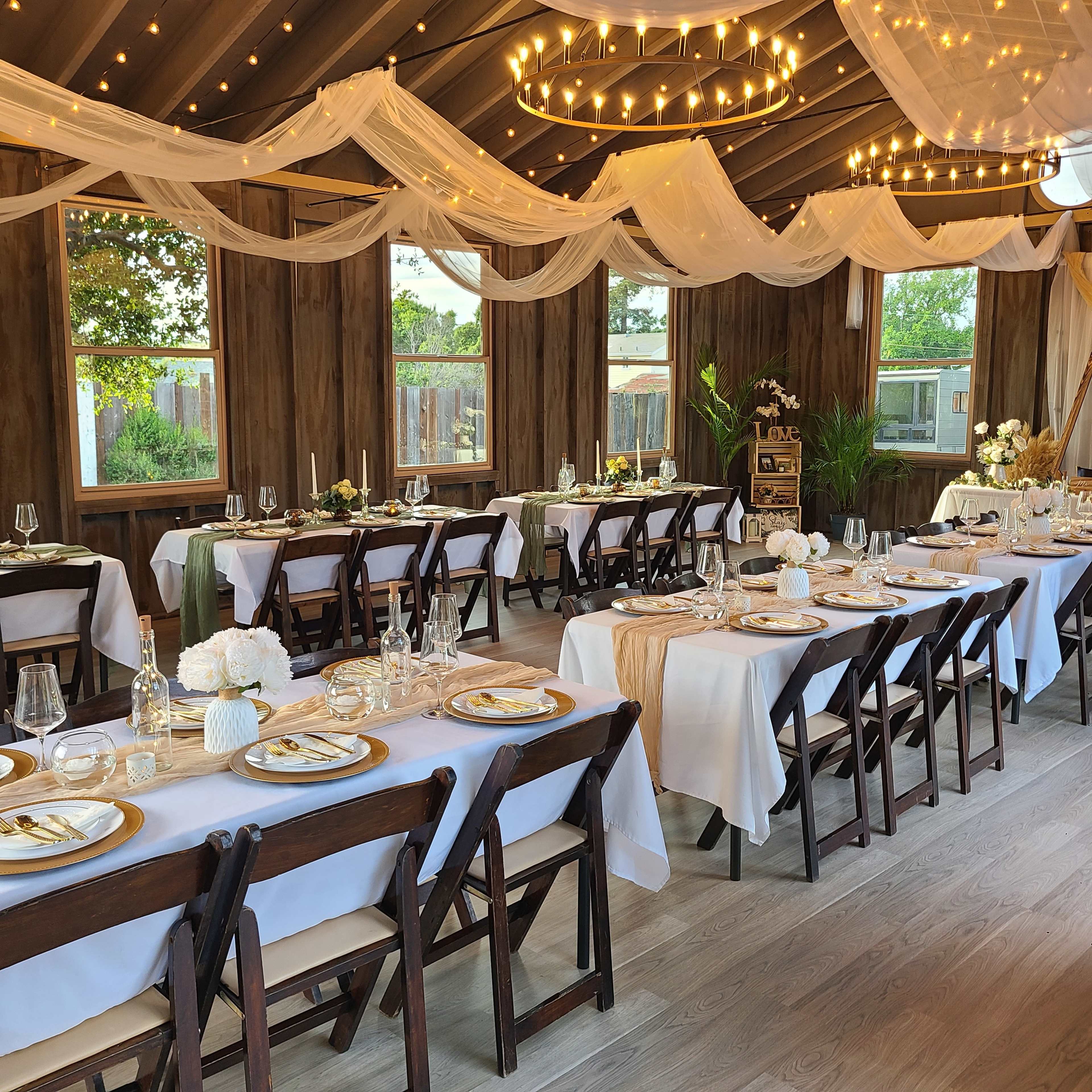 A beautifully arranged dining area features multiple tables set with white tablecloths, elegant dishware, and decorative floral centerpieces under soft, draped fabric and string lights.