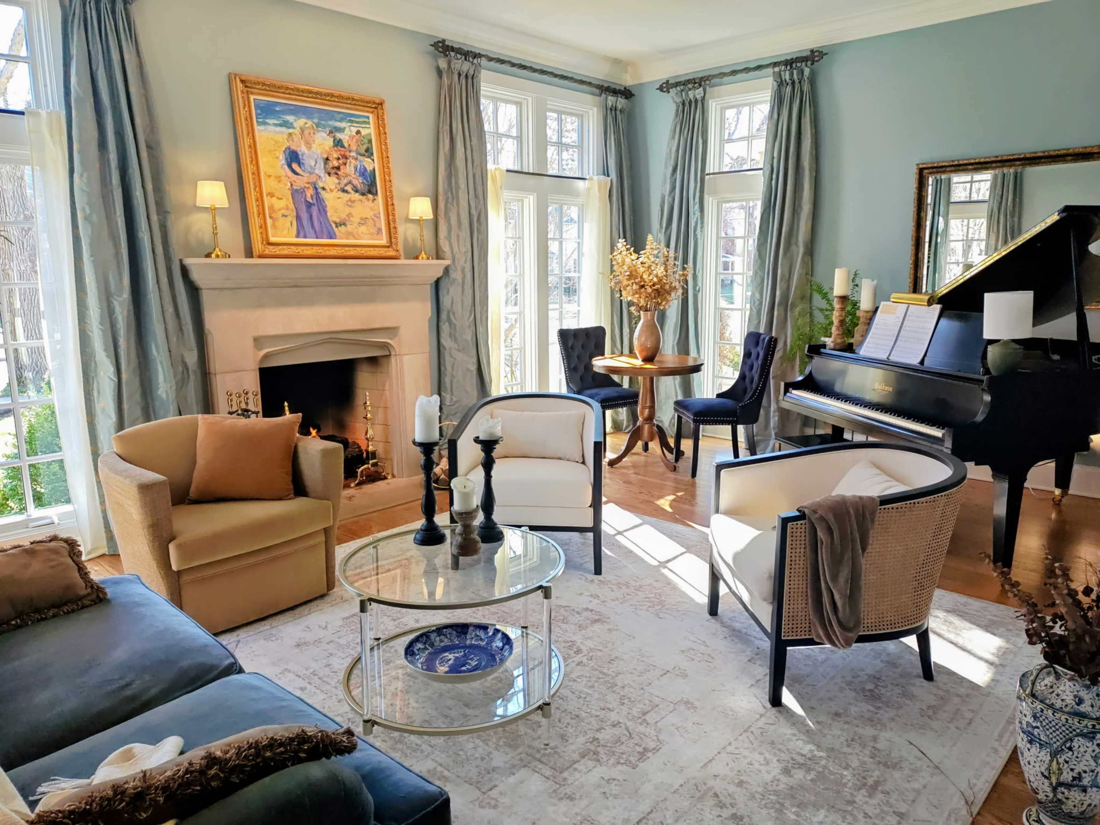 The room features a piano, a round table, and various seating arrangements, all accented by natural light streaming through large windows.