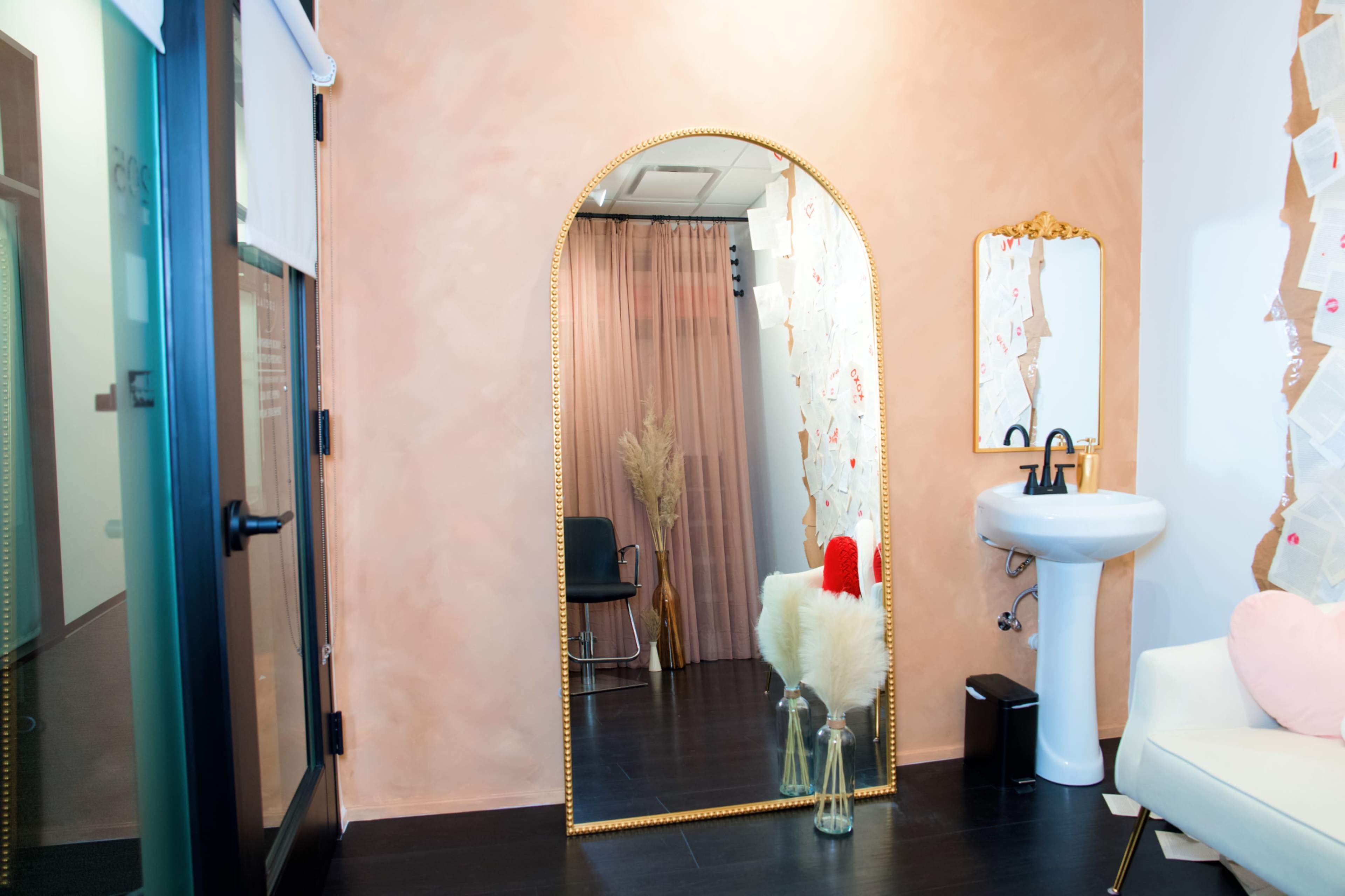 A large, arched mirror reflects a pink wall and a small white sink in a decoratively arranged room.