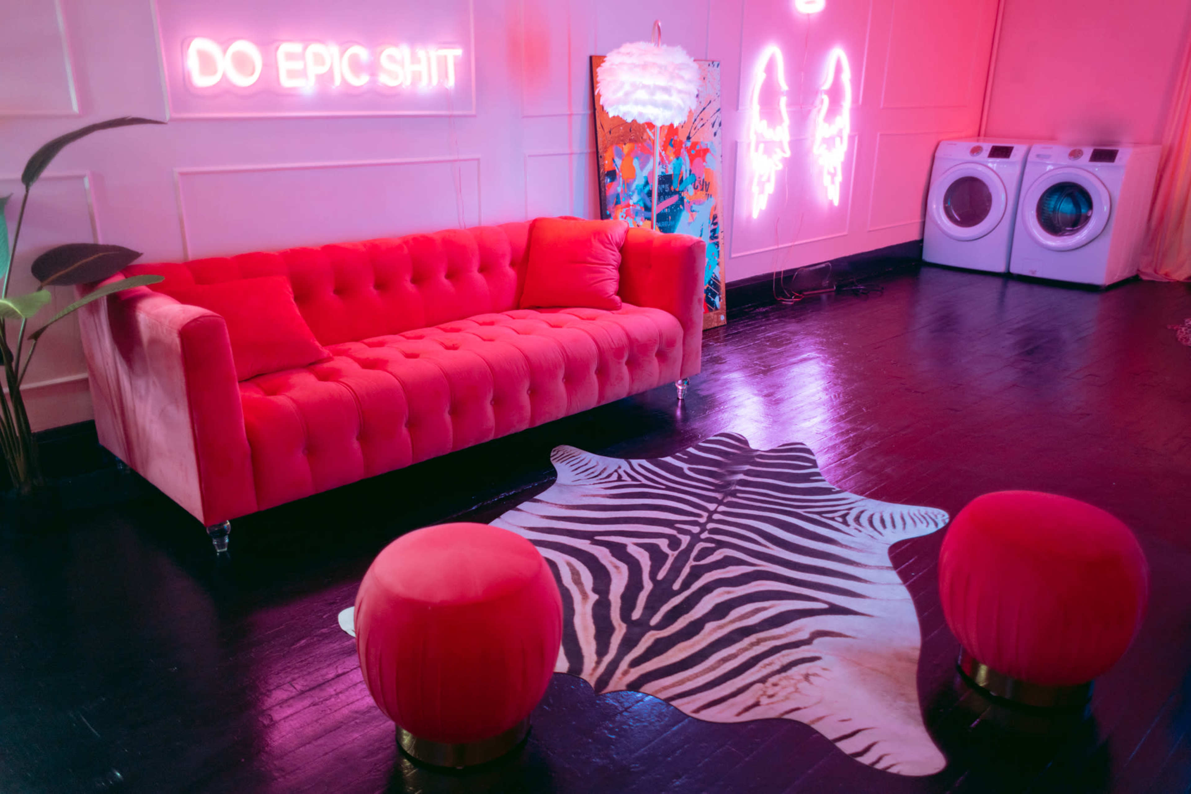 A brightly colored room featuring a pink tufted sofa, a zebra print rug, two round stools, and a wall with neon lights.
