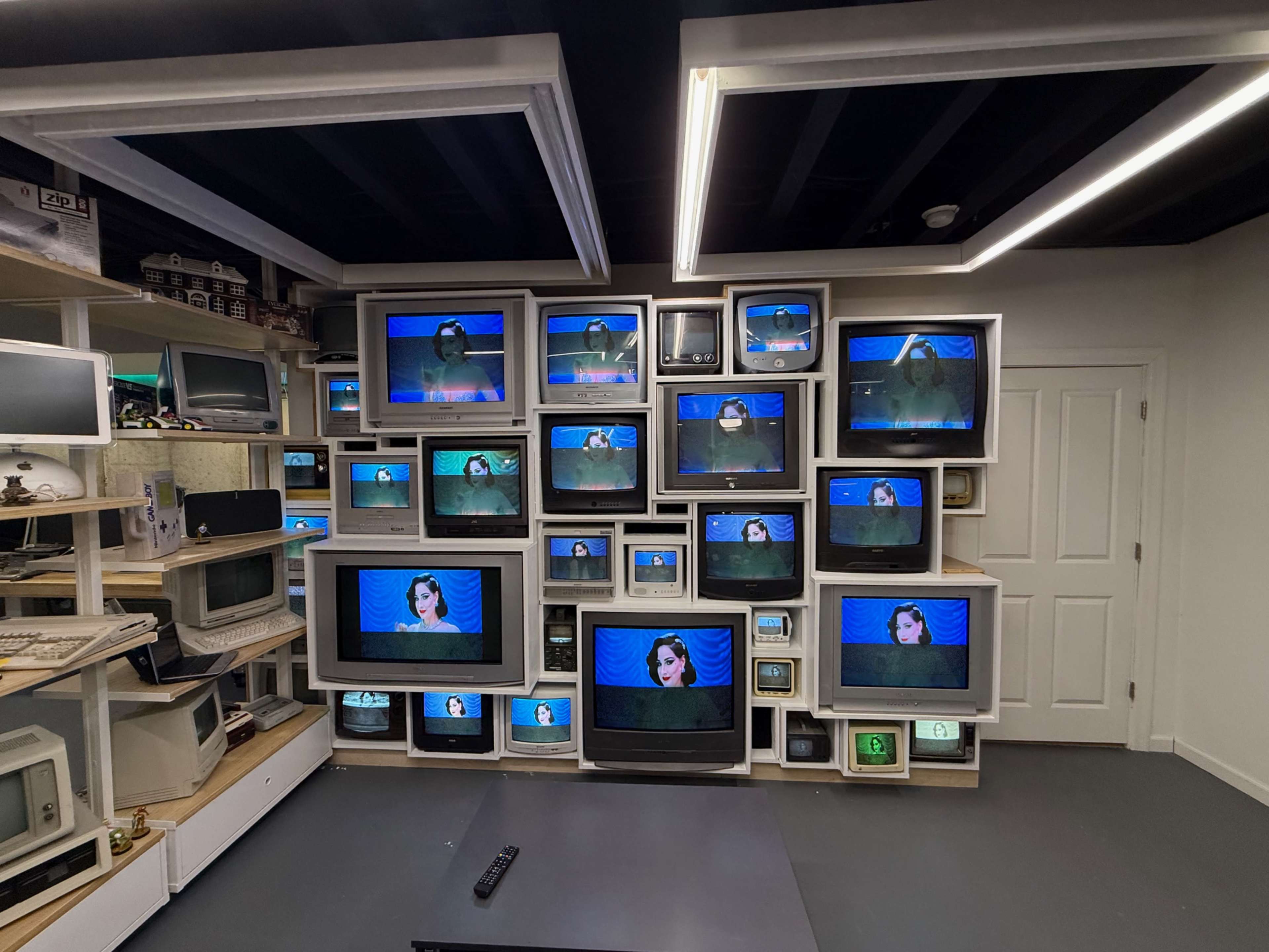The image shows a wall filled with various old television sets and computer monitors, all displaying a woman on a blue background.