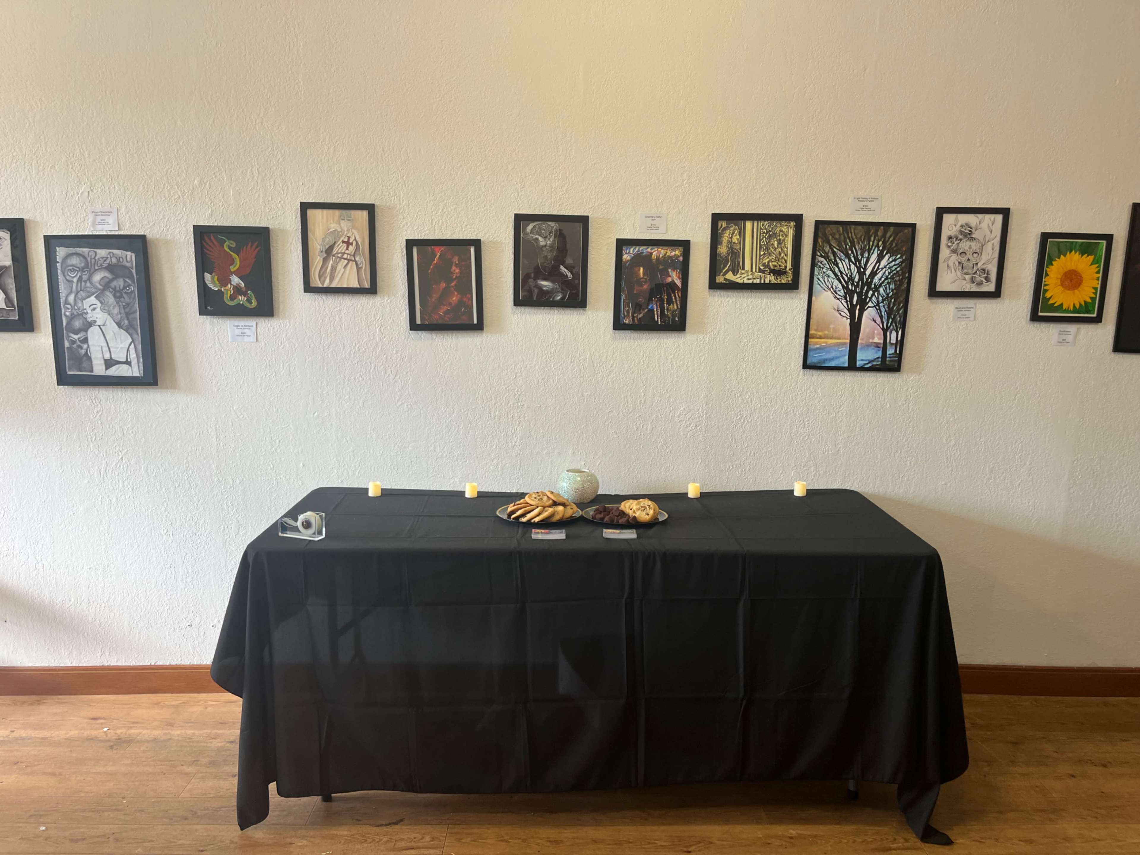The image shows a gallery wall displaying framed artworks alongside a black table with candles and a decorative bowl set in front of it.