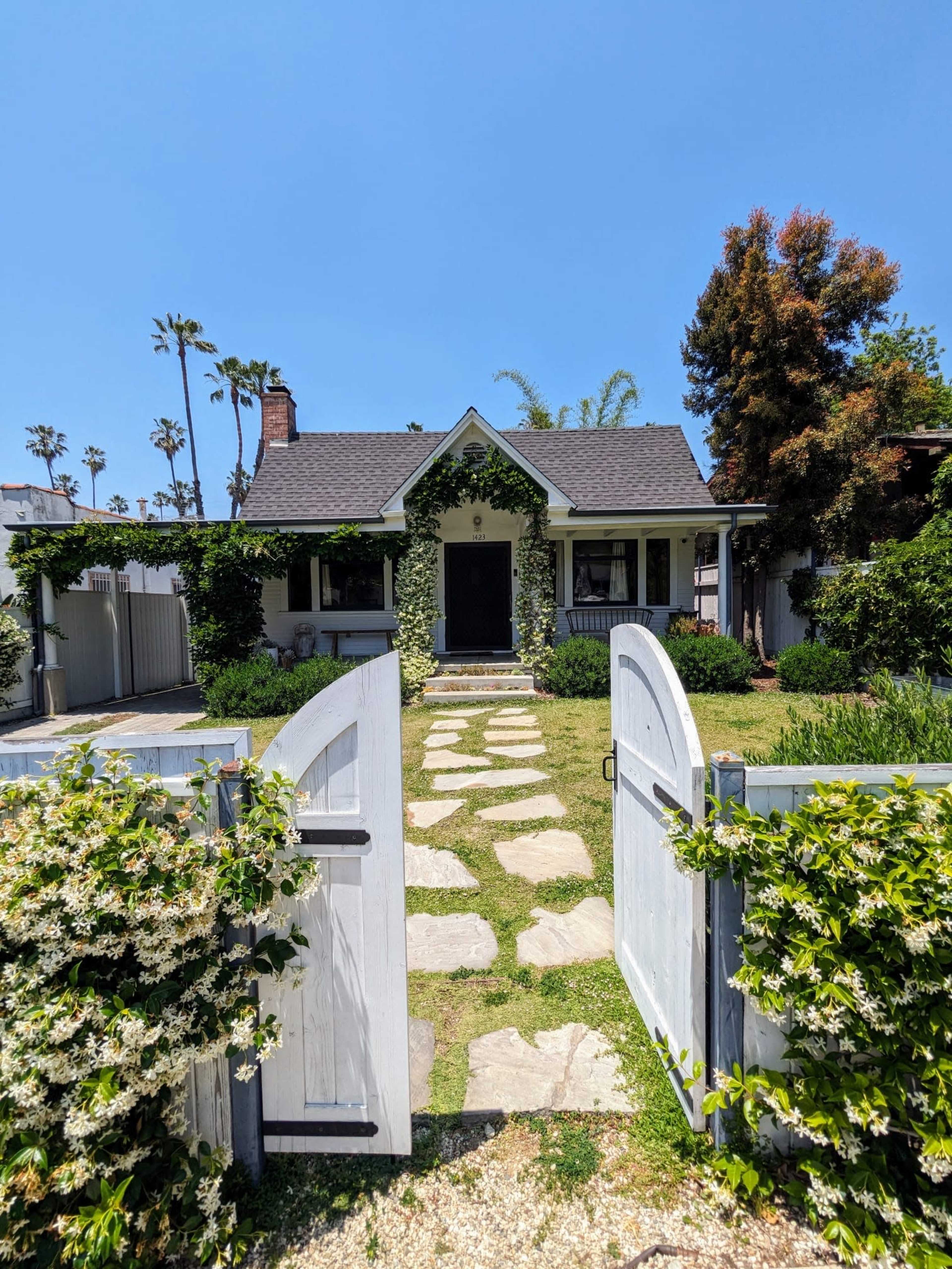 A white picket fence with open gates leads to a bungalow surrounded by greenery and a stone pathway.