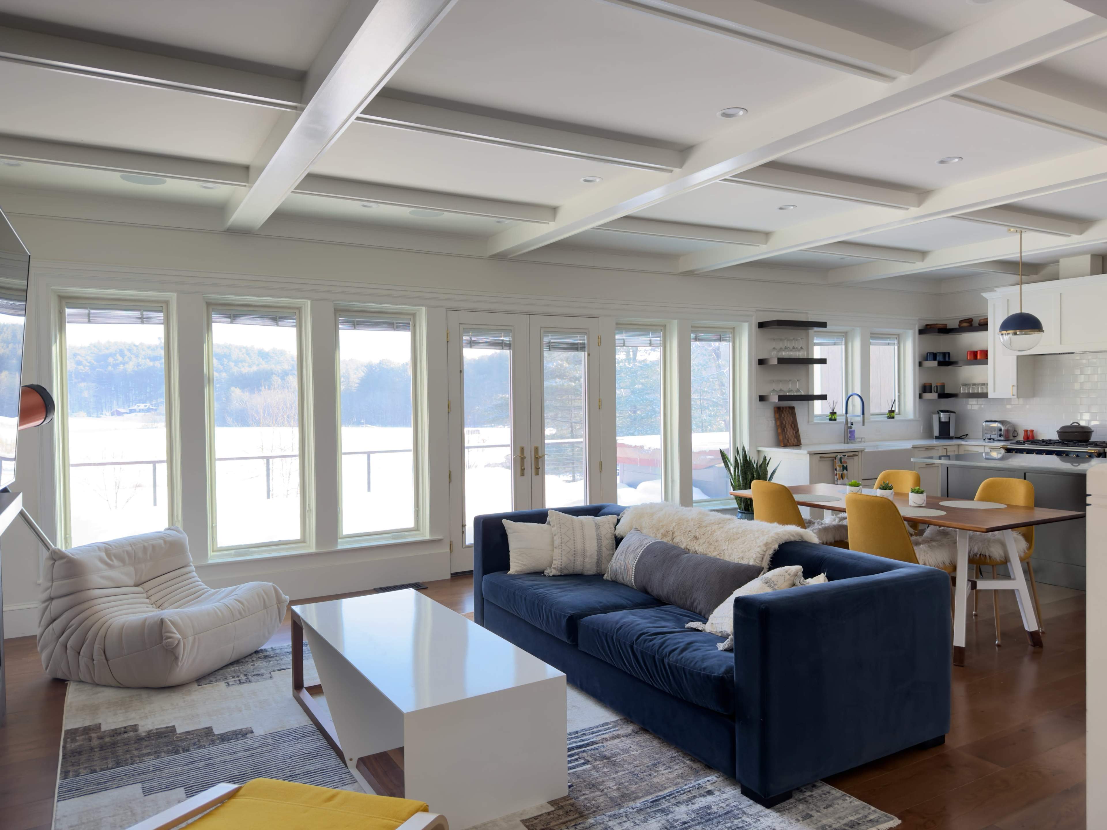The image shows a modern living room with large windows overlooking a scenic view, featuring a blue sofa, a white coffee table, and a dining area with yellow chairs.