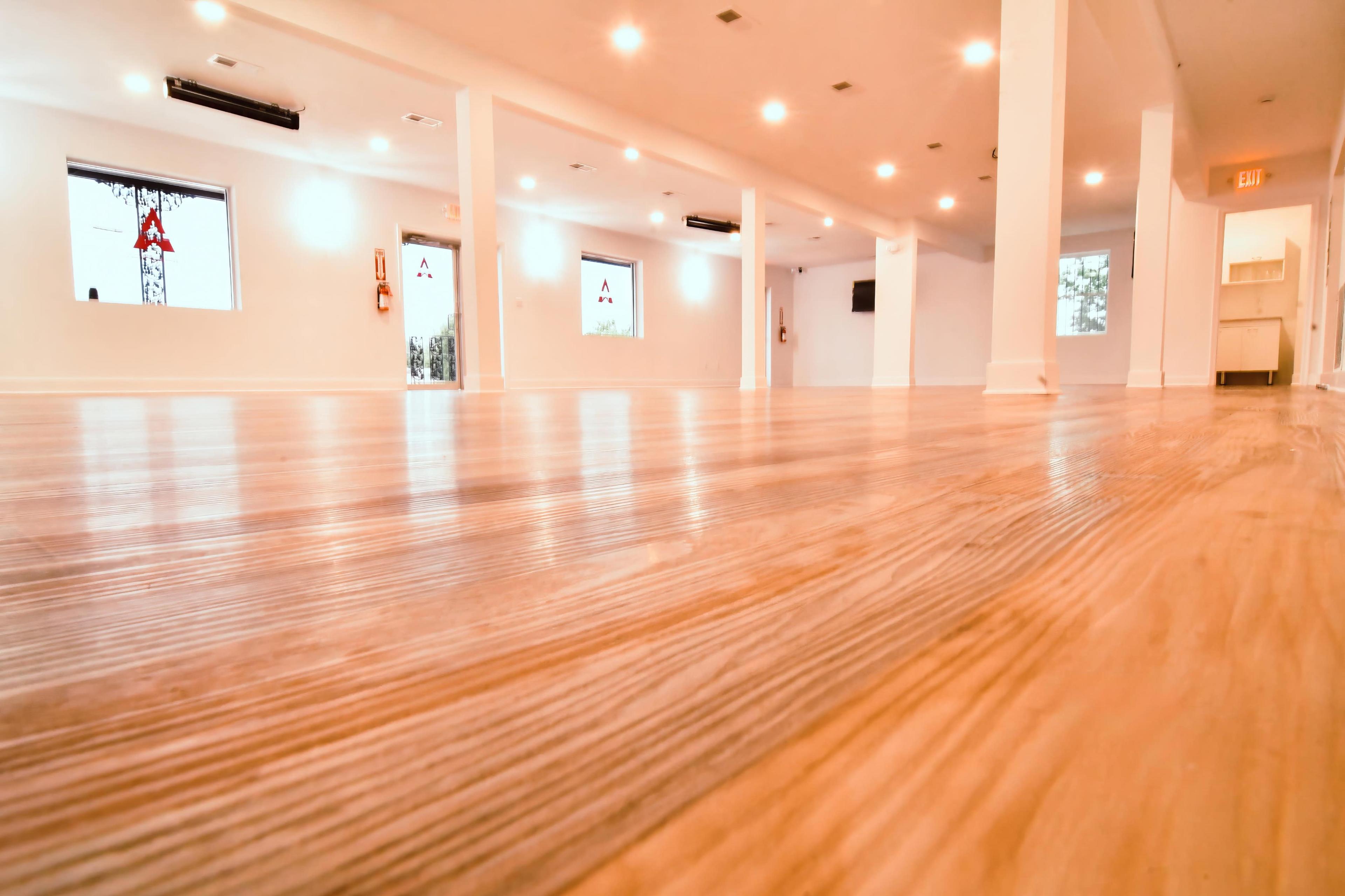 The image shows a spacious, well-lit indoor area with polished wooden floors and white walls, featuring several windows and lighting fixtures.