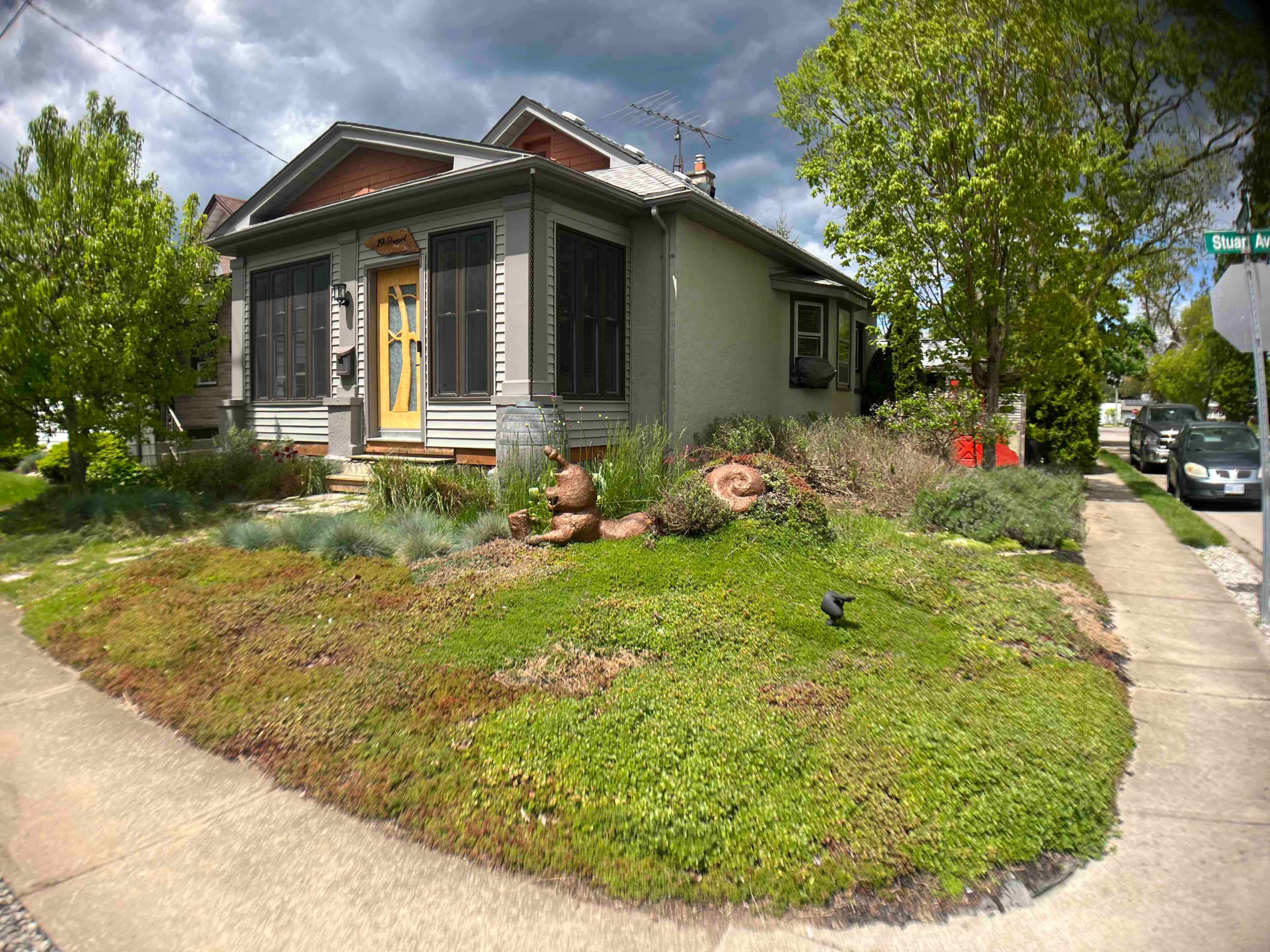 A house with a yellow front door is surrounded by a well-maintained garden featuring shrubs and decorative stones along a sidewalk.