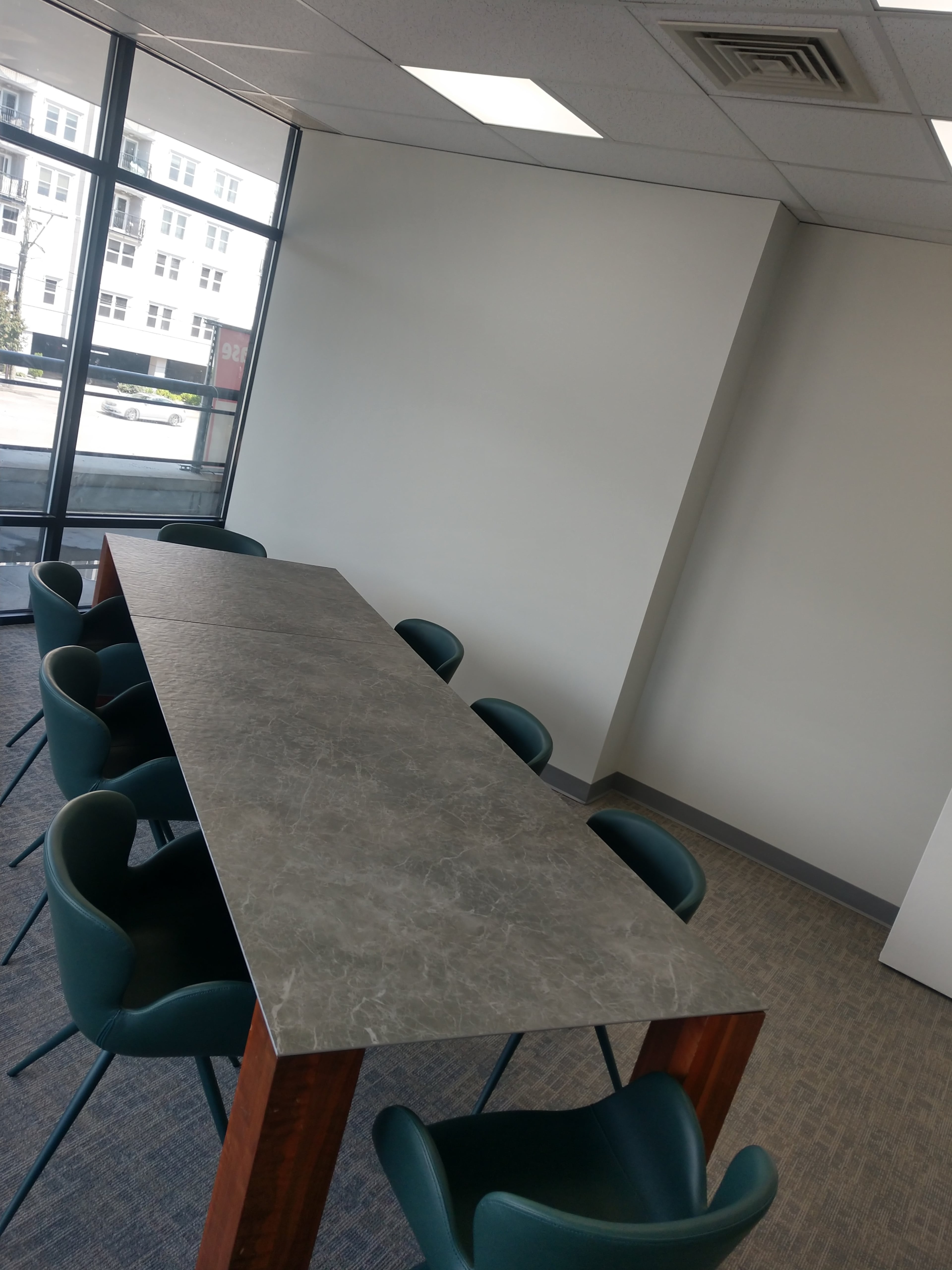 A long, rectangular table made of gray stone is surrounded by eight green chairs in a well-lit meeting room with large windows.